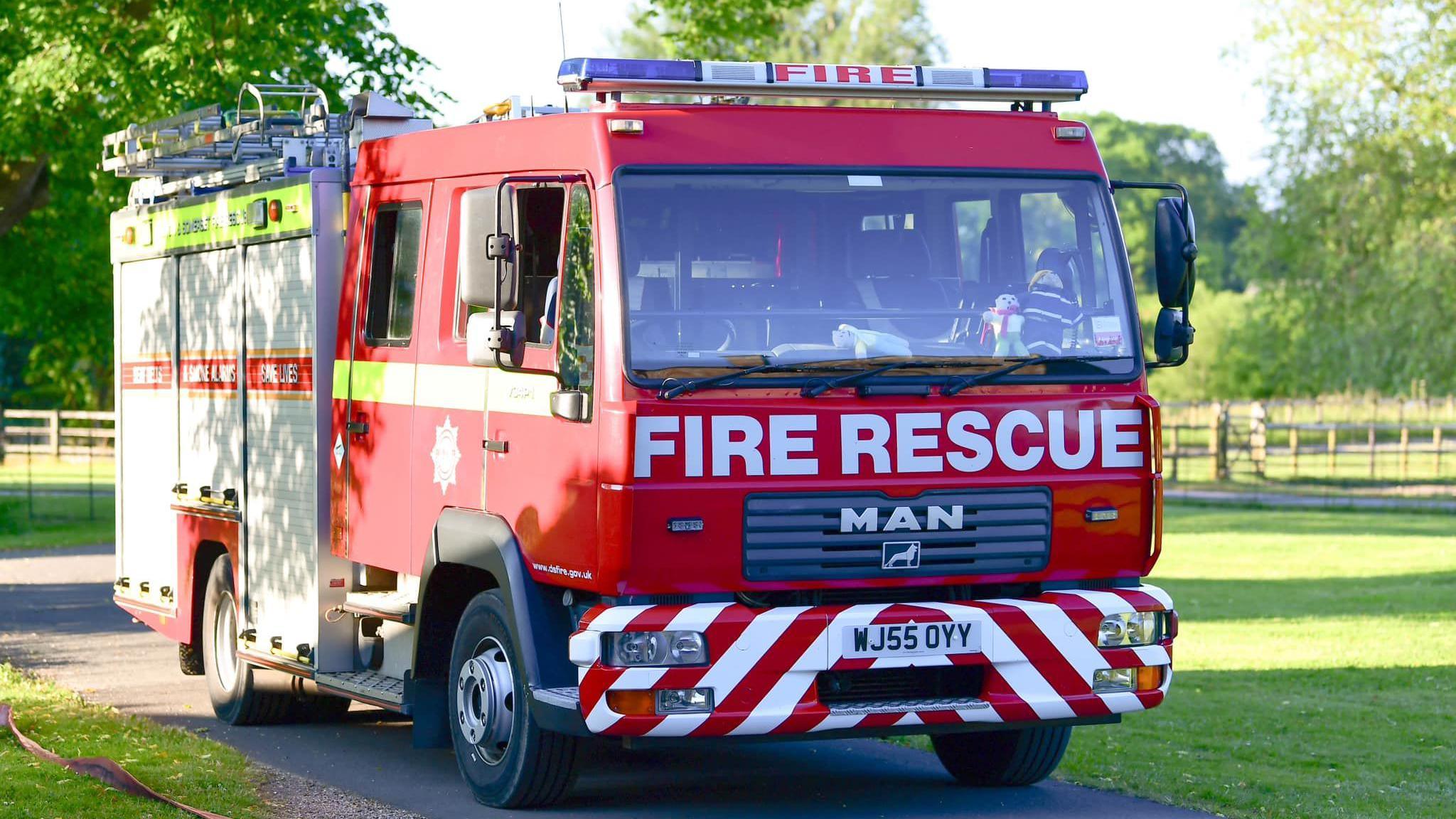 A red fire engine on a path surrounded by grass lawns and trees on a sunny day. Knitted toys including one of a firefighter are on the dashboard.