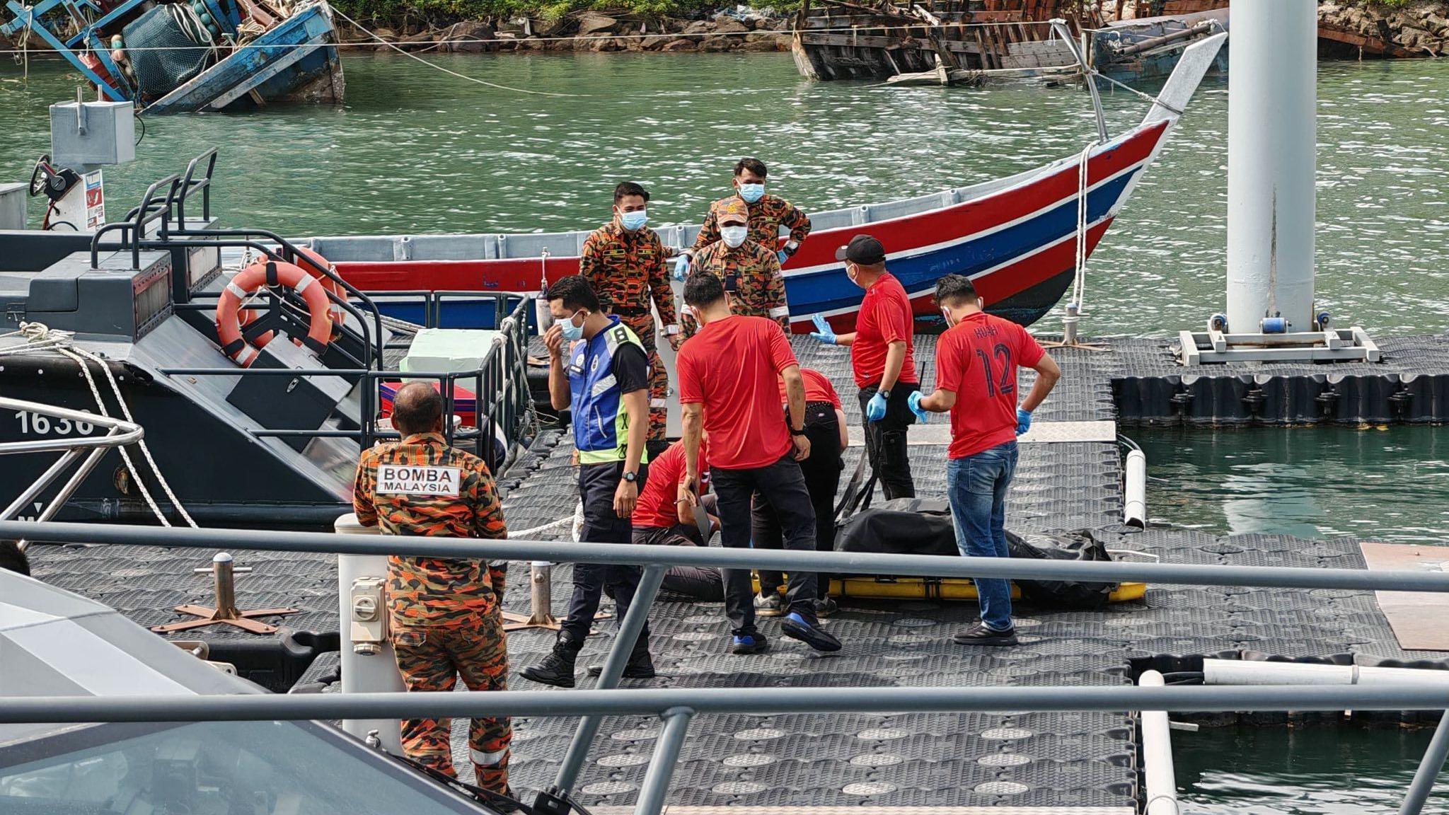 Men in red shirts and green/orange camouflage attire standing around a black body bag lying on a stretcher.