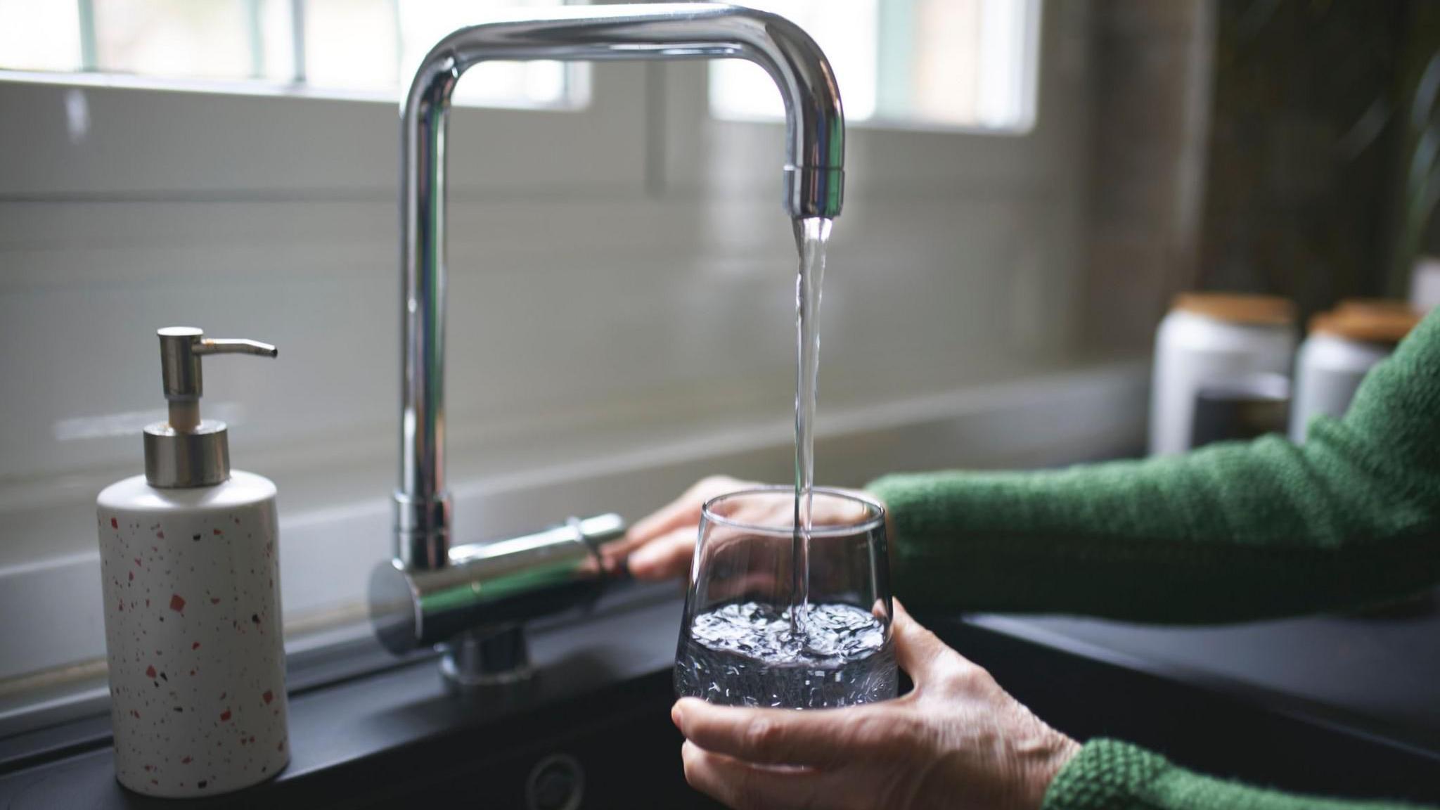 A woman is pictured filling a glass with water from a tap. The basin and soap dispenser are also visible.