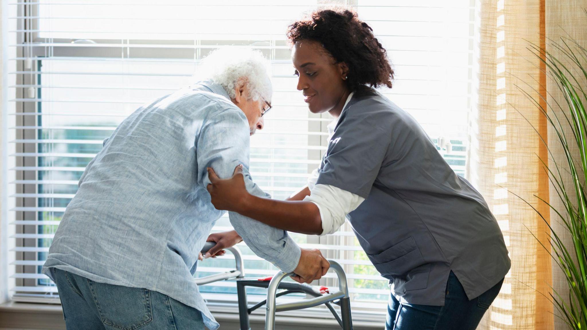 A care home worker helps support an elderly man leaning on a walking frame