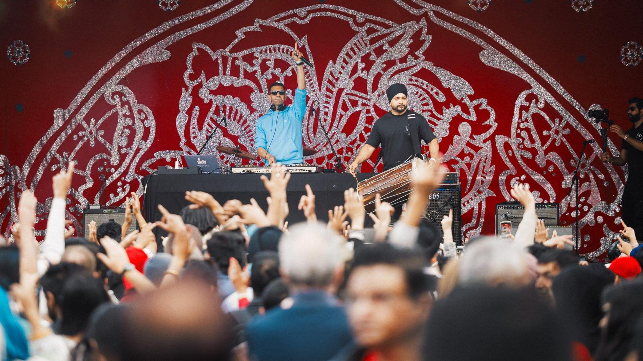A DJ and musician stand onstage in front of a cheering crowd - the DJ is wearing a blue top and has one hand raised in the air while at the decks