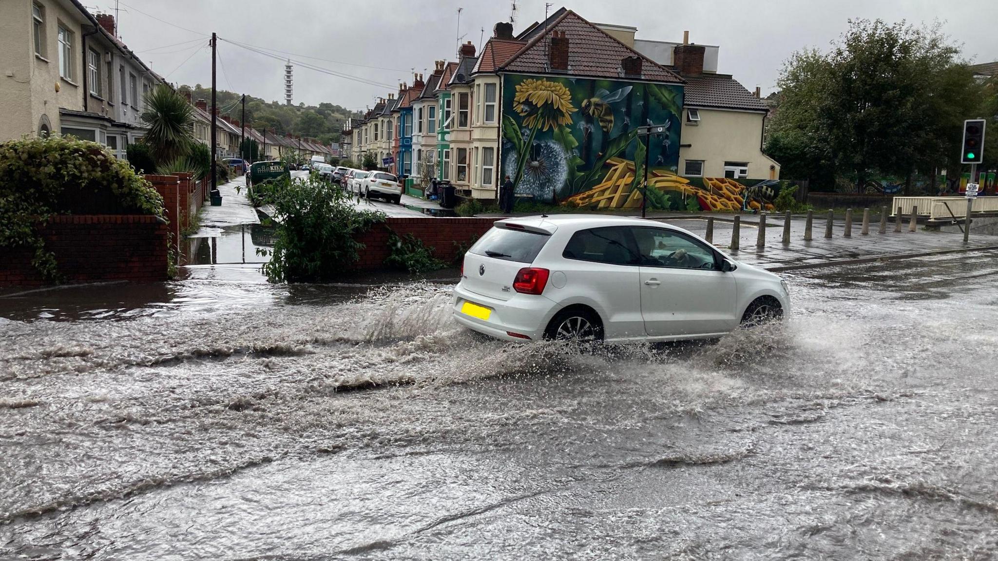 A white car drives through floodwater close to the M32 in Eastville, Bristol. A row of terraced houses is visible, as is the Lockleaze Tower in the background