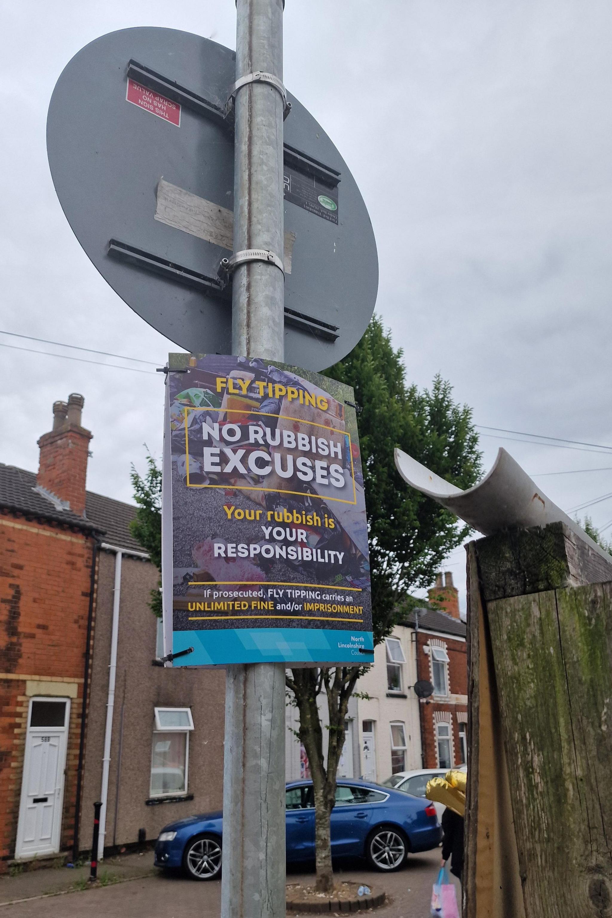 A council sign on a post warns against fly-tipping and reads "No rubbish excuses". There is a fence to the side and houses behind it. There are also vehicles behind it.