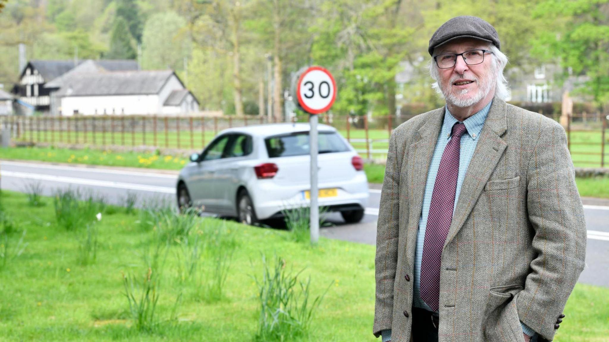 Councillor Peter Thornton standing on a Patterdale Road. He has grey hair and a grey beard, and is wearing glasses, a dark flat cap, tweed blazer and a shirt and tie. Behind him, a car is travelling along the road and a 30mph sign is visible.