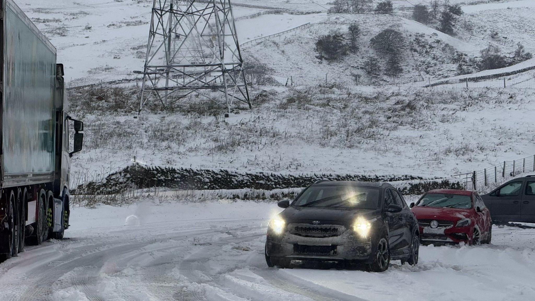 Three cars struggling to drive on the A6 heading up hill. Two cars have been forced to drive to the side of the road. A third car behind them is facing across the road towards a field.
A lorry, to the right, is driving down hill.