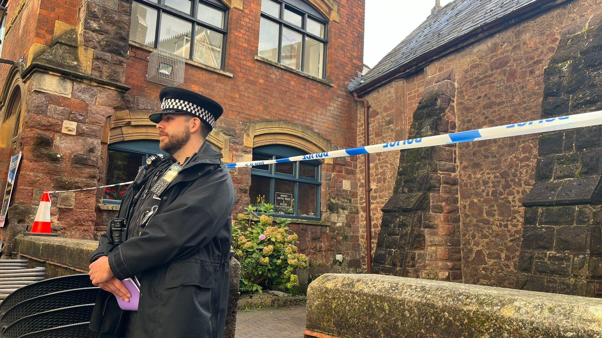 A male police officer wearing a hat and black coat stood in front of blue and white police tape. The officer is also holding a purple notebook in his left hand. The tape is surrounding a brown building on Catherine Street in Exeter. 