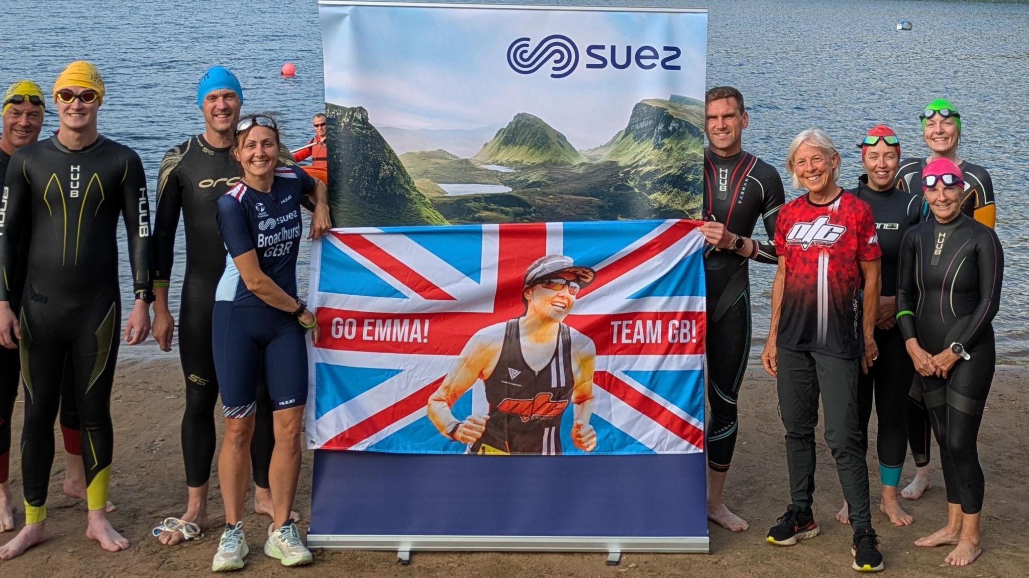A woman wearing a dark blue Team GB outfit, holds a union flag which has an image of her running, with "Go Emma" and "Team GB" in white lettering. Other people dressed in wetsuits are standing around her. A lake is visible in the background.