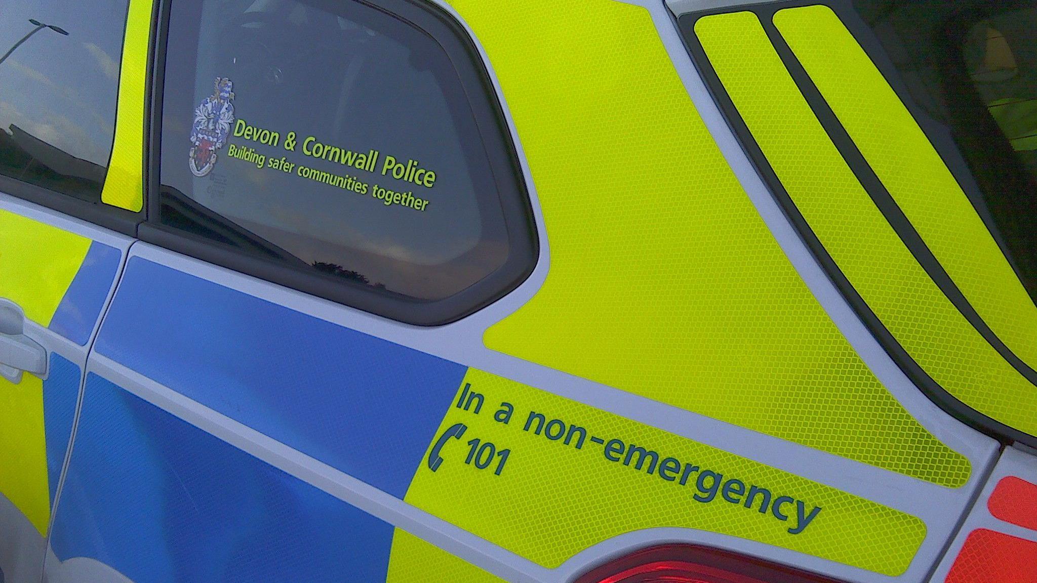 The side of a yellow and blue police vehicle. On the back window, there are the words 'Devon and Cornwall Police' in yellow writing with a badge to the left of it.  On the back of the police car is the words 'In a non-emergency' call 101. 