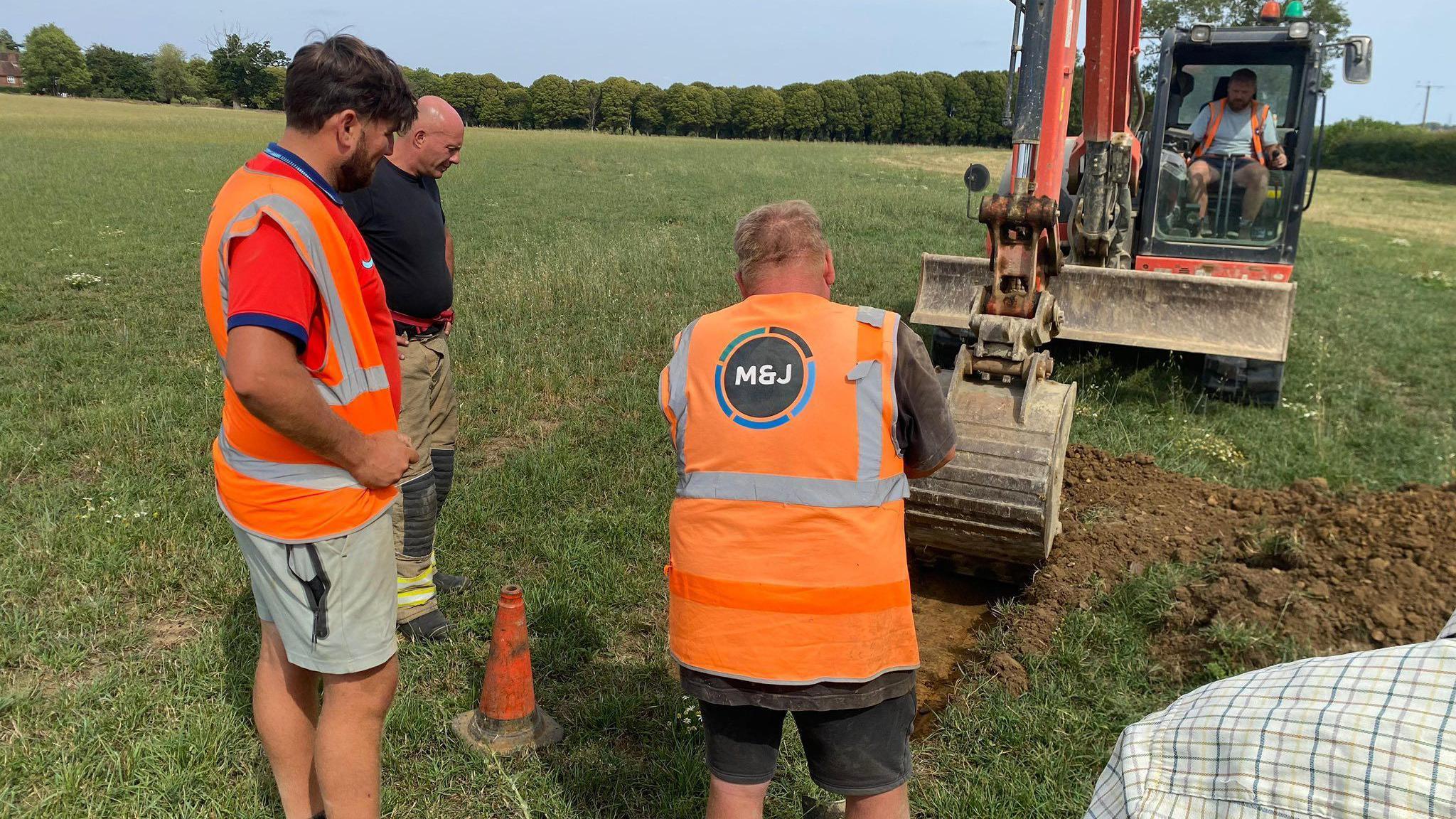 Three people in hi-vis watch on as a man in a digger carves out a trench in the middle of a green field.