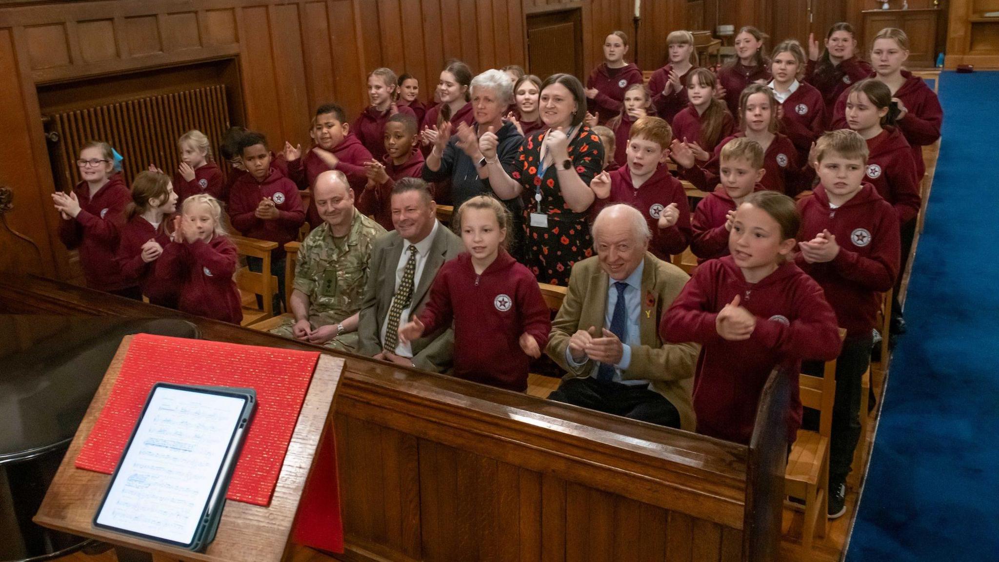 Children sit singing in church pews, encouraged by two men and two women sat among them.