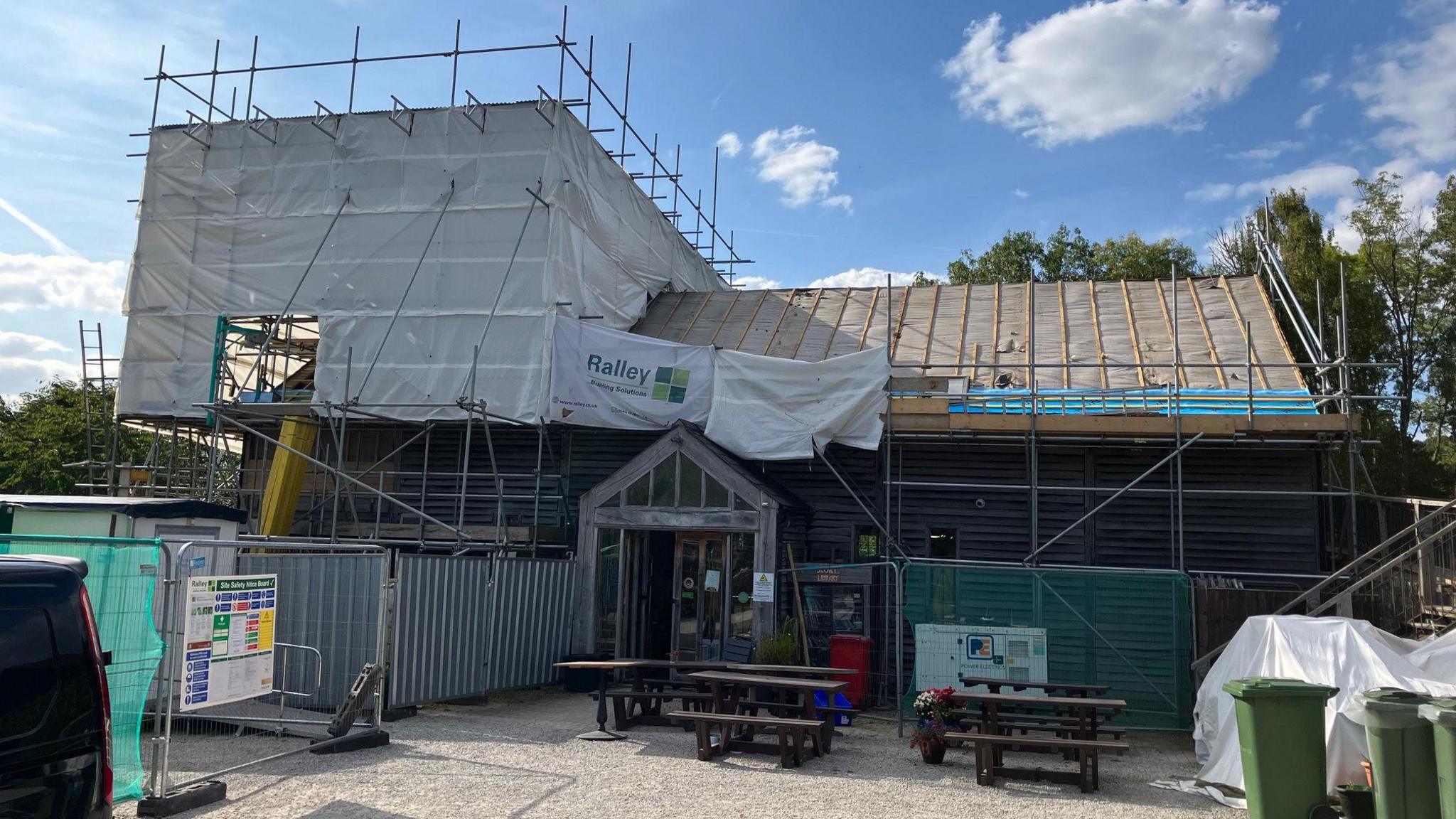 The exterior of the café and shop, taken on a sunny day in August when the whole building was under construction to repair the fire and water damage. There is scaffolding and plastic sheeting surrounding the building. The roof tiles have been taken down and you can see the insulation beneath. Outside the entrance there is metal heras fencing, green bins, and wooden picnic benches stacked in a pile.