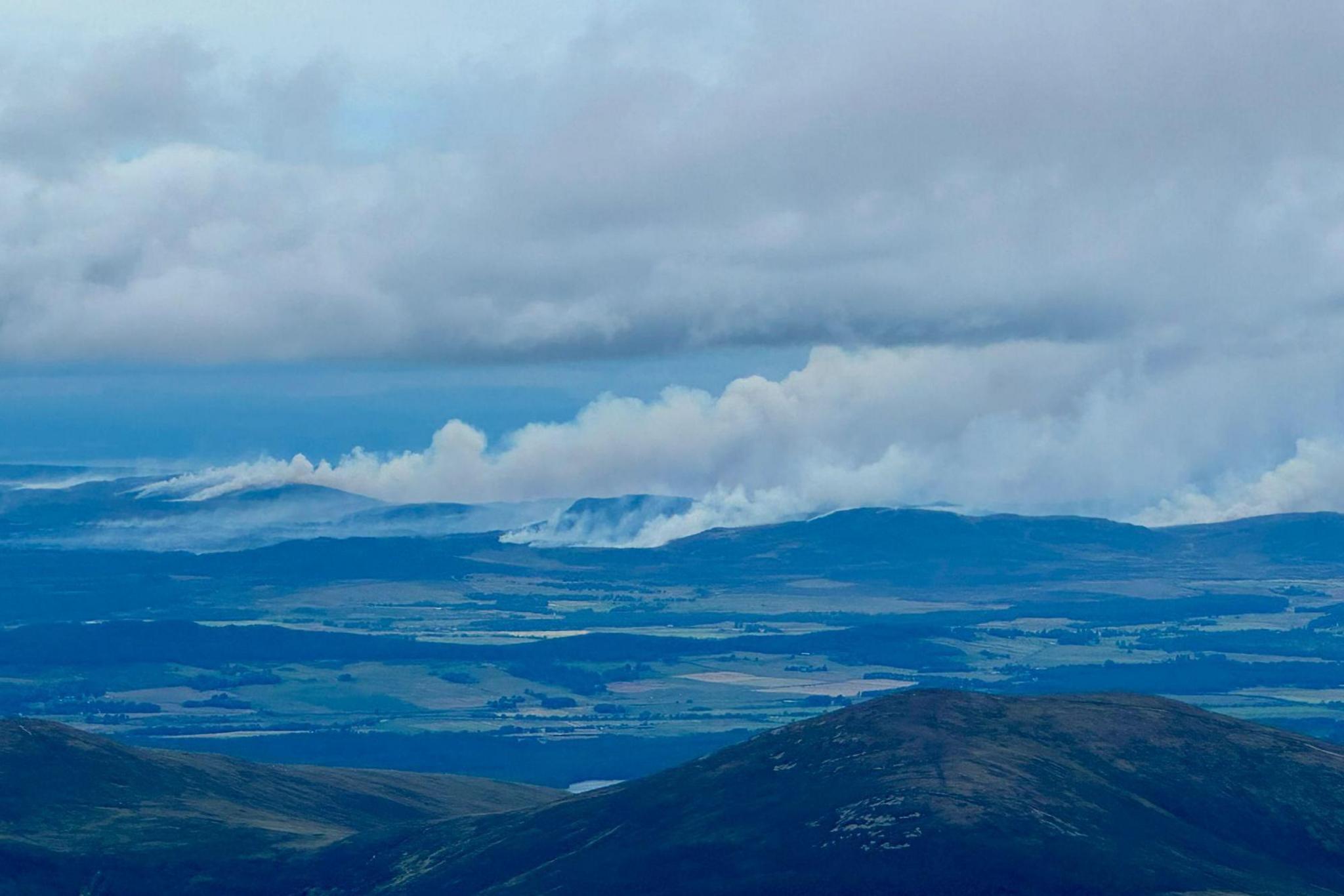 A view across hills, fields and forests towards thick smoke rising from several wildfires.