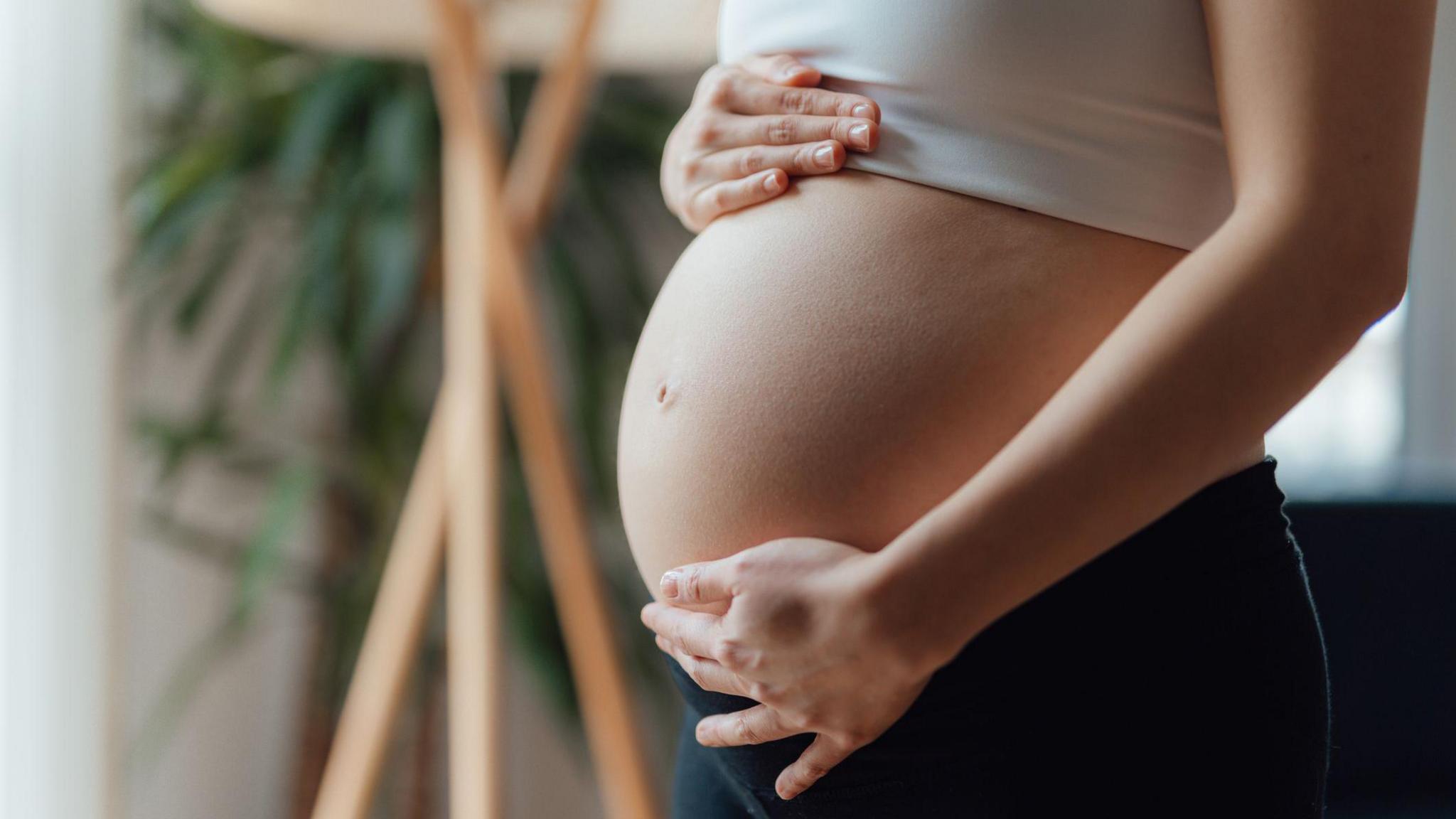 A pregnant woman places her hand on the top and bottom of her baby bump. She is wearing a white cropped top and her pregnant belly is centre of the frame.She is wearing black leggings and is in a living room.