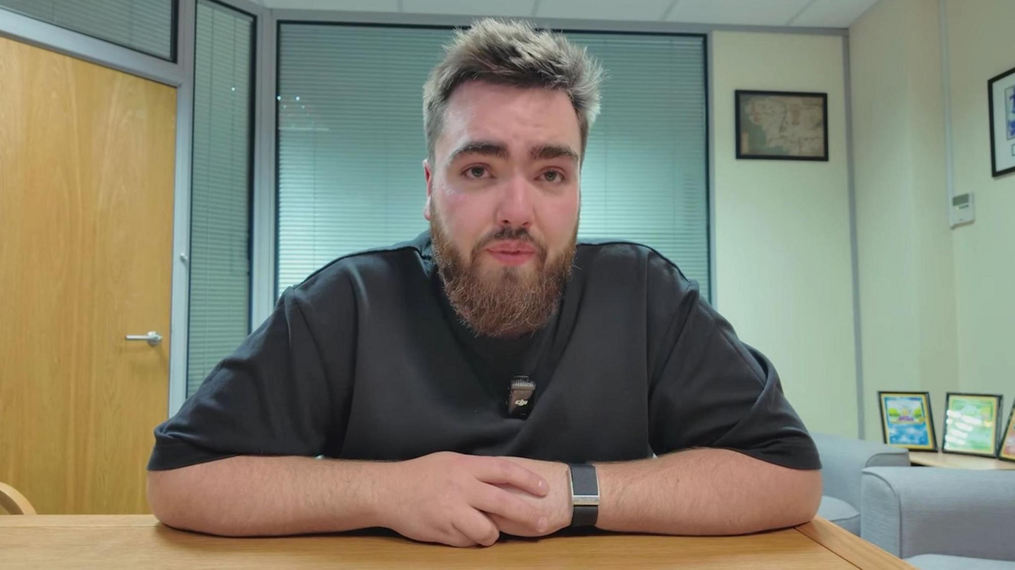 Randolph is sat with his arms crossed. He has a brown beard and brown hair, and is wearing a black t-shirt. On office is behind him.