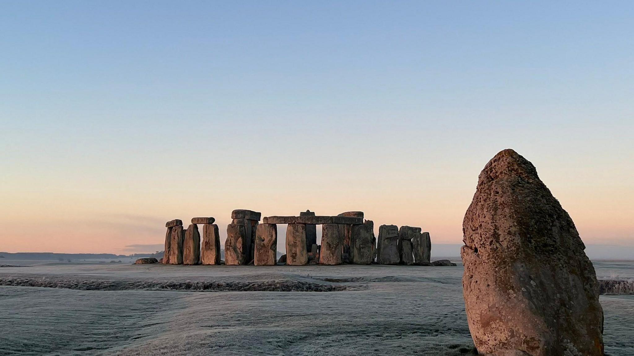 A picture of Stonehenge taken from afar. The top of the sky is blue and fades into an orange at the horizon. The grass surrounding the monument is covered in frost.