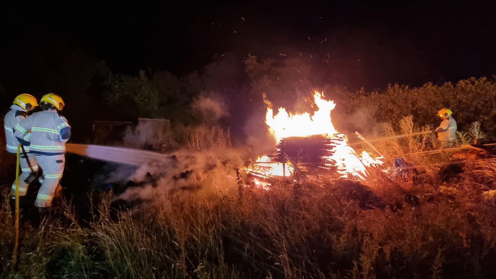 Two firefighters holding a large hose reel jet, which is spraying water at a stack of wood in a field with long grass. There is another firefighter in the background spraying another area. The fire has bright orange flames and sparks flying from above