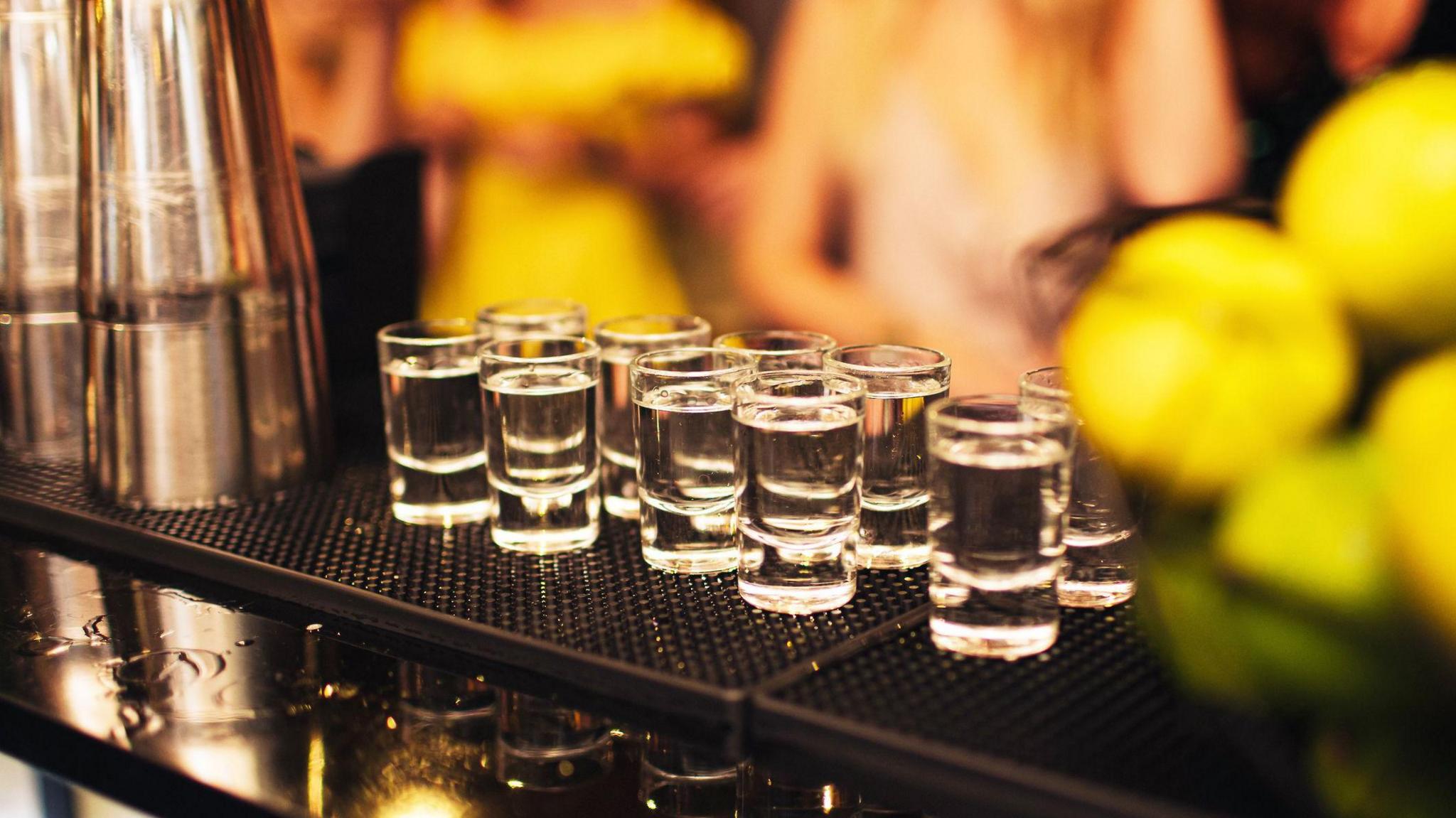 Ten shot glasses filled with clear liquid lined up on a bar next to a cocktail shaker. There are people blurred in the background.