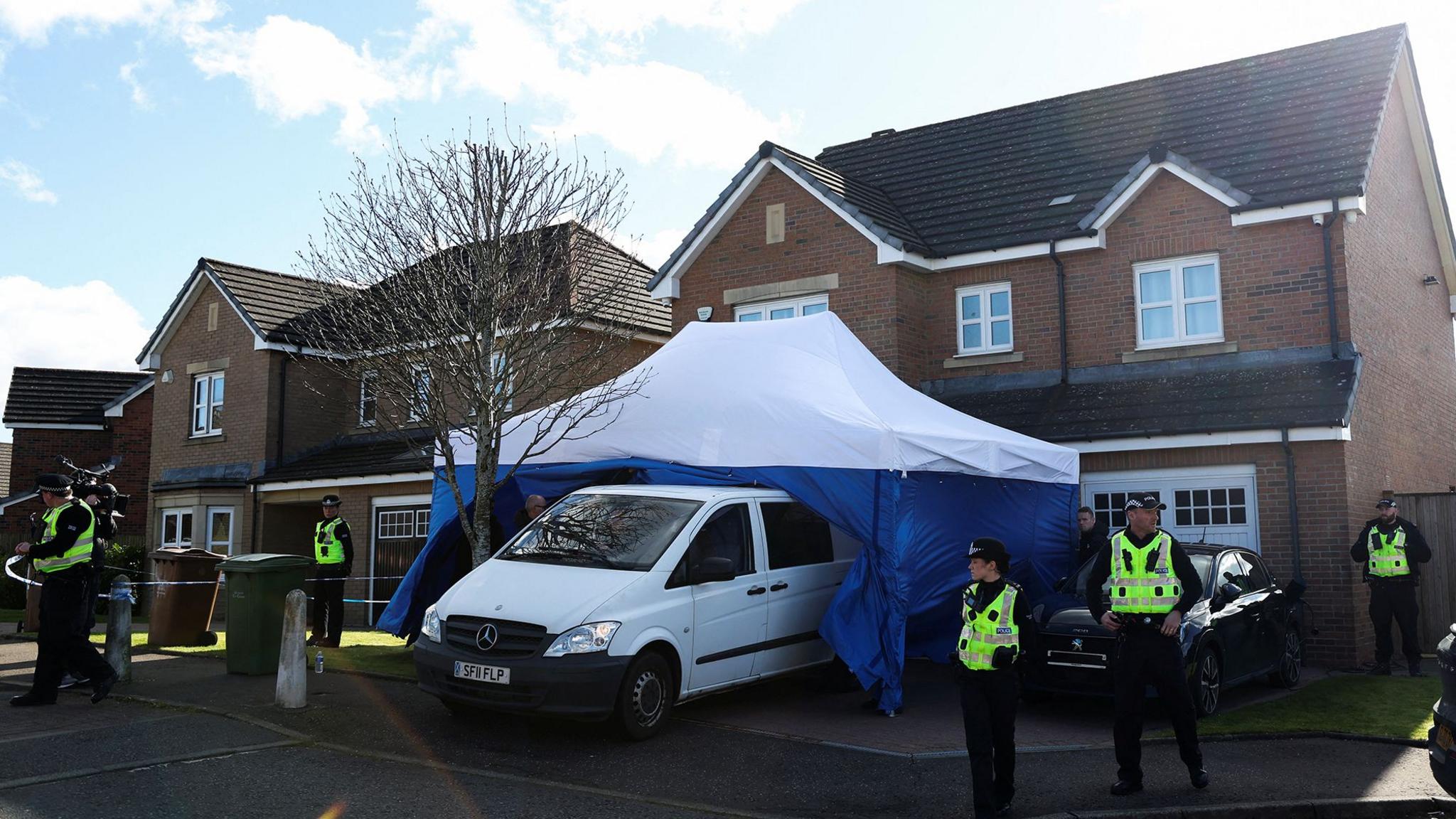 Police officers and a large blue an white police tent outside a modern red brick house