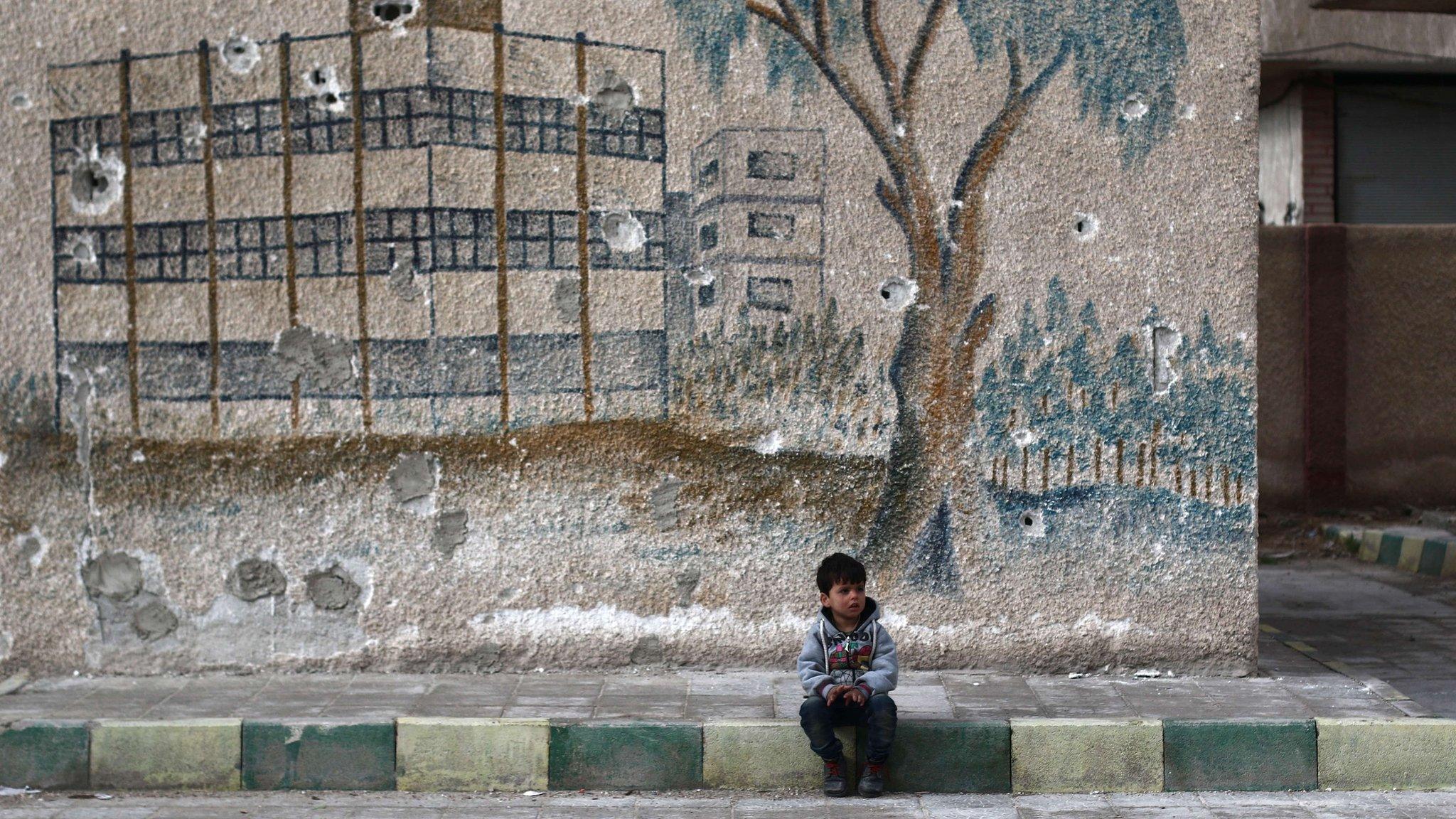 A Syrian child sits in front of a mural covered in bullet holes on the wall of a former school in the rebel-held region of Eastern Ghouta, on the outskirts of the Syrian capital Damascus, on January 5, 2016.