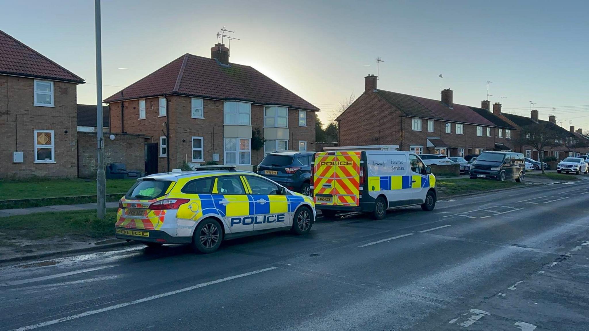 A police car and police forensic van are parked at the side of Hawthorn Drive. Houses can be seen to the left of them with other vehicles parked on a grass verge.