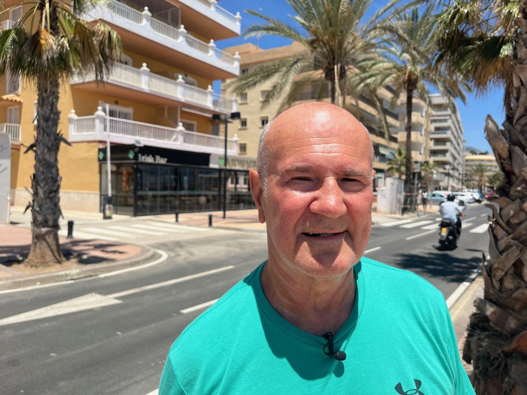 Robert Sorman smiles at the camera - he is standing on a street lined with palm trees and is wearing a turquoise Under Armour t-shirt. He is mostly bald with some closely shaved white hair.