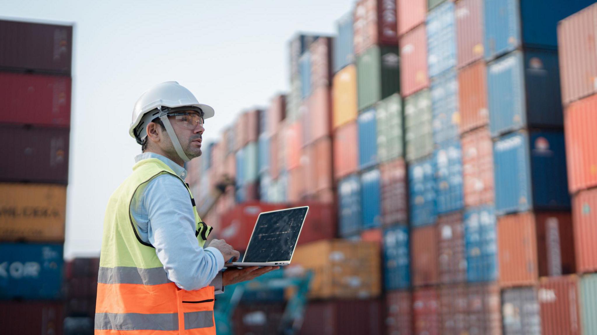 A port worker using a laptop and standing close to stacks of shipping containers.   The man is wearing a high-vis vest, a safety helmet and safety glasses. 