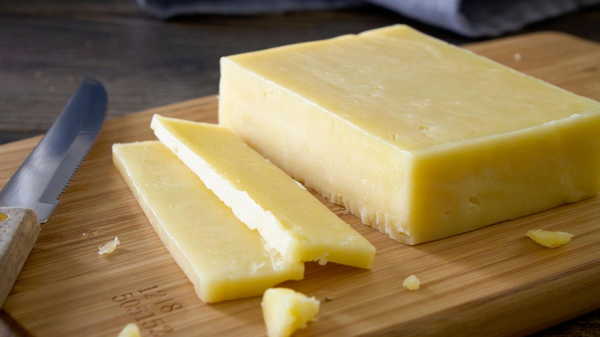 A block of cheese on a chopping board with two slices taken out of it. A knife sits beside the cheese, also on the chopping board.