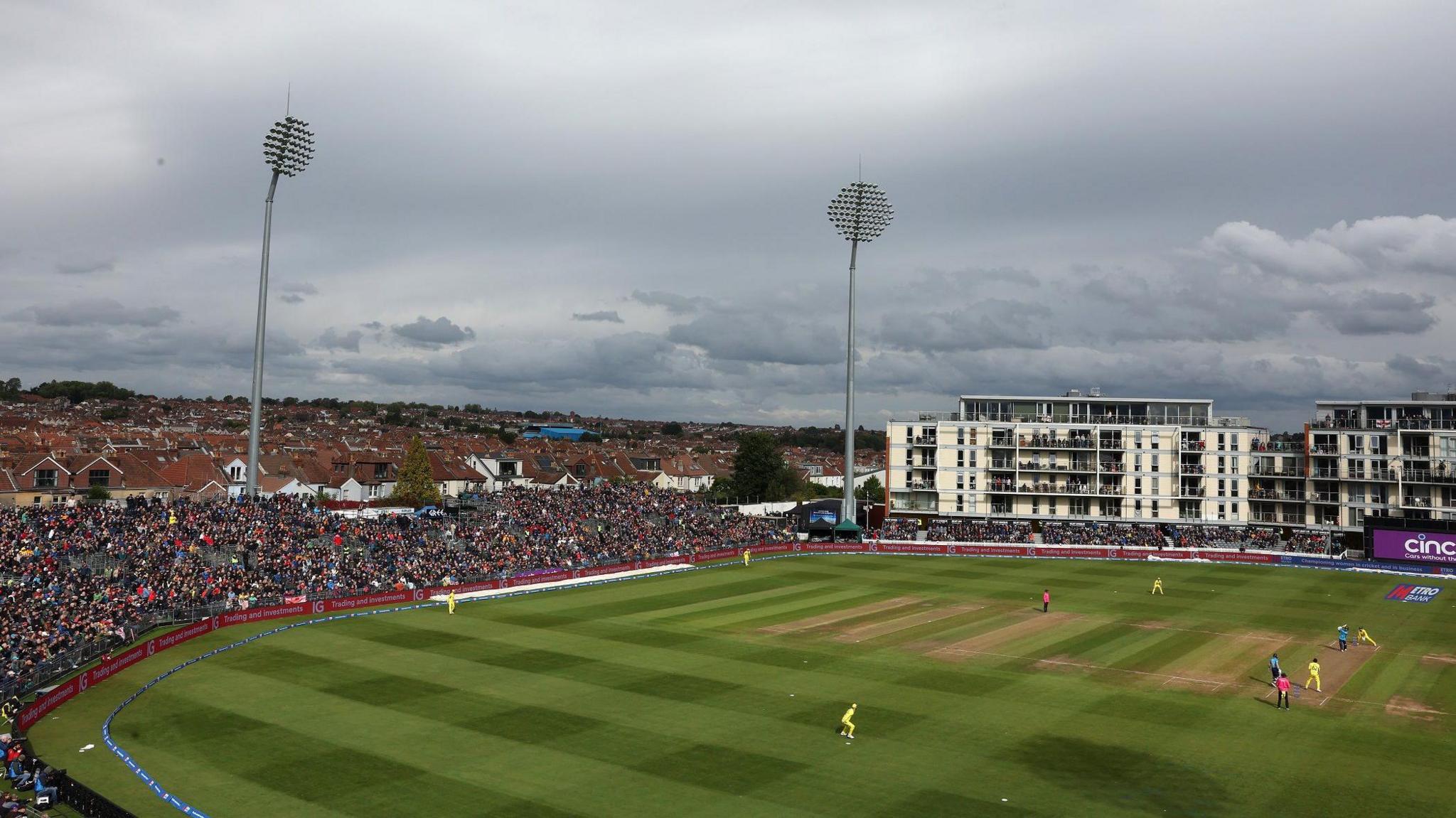 A wide shot of the county cricket ground in Bristol as England played Australia in a one-day international. Australia are fielding in their yellow kit and the full grandstands and housing estates are visible in the background, along with the stadium's large floodlights