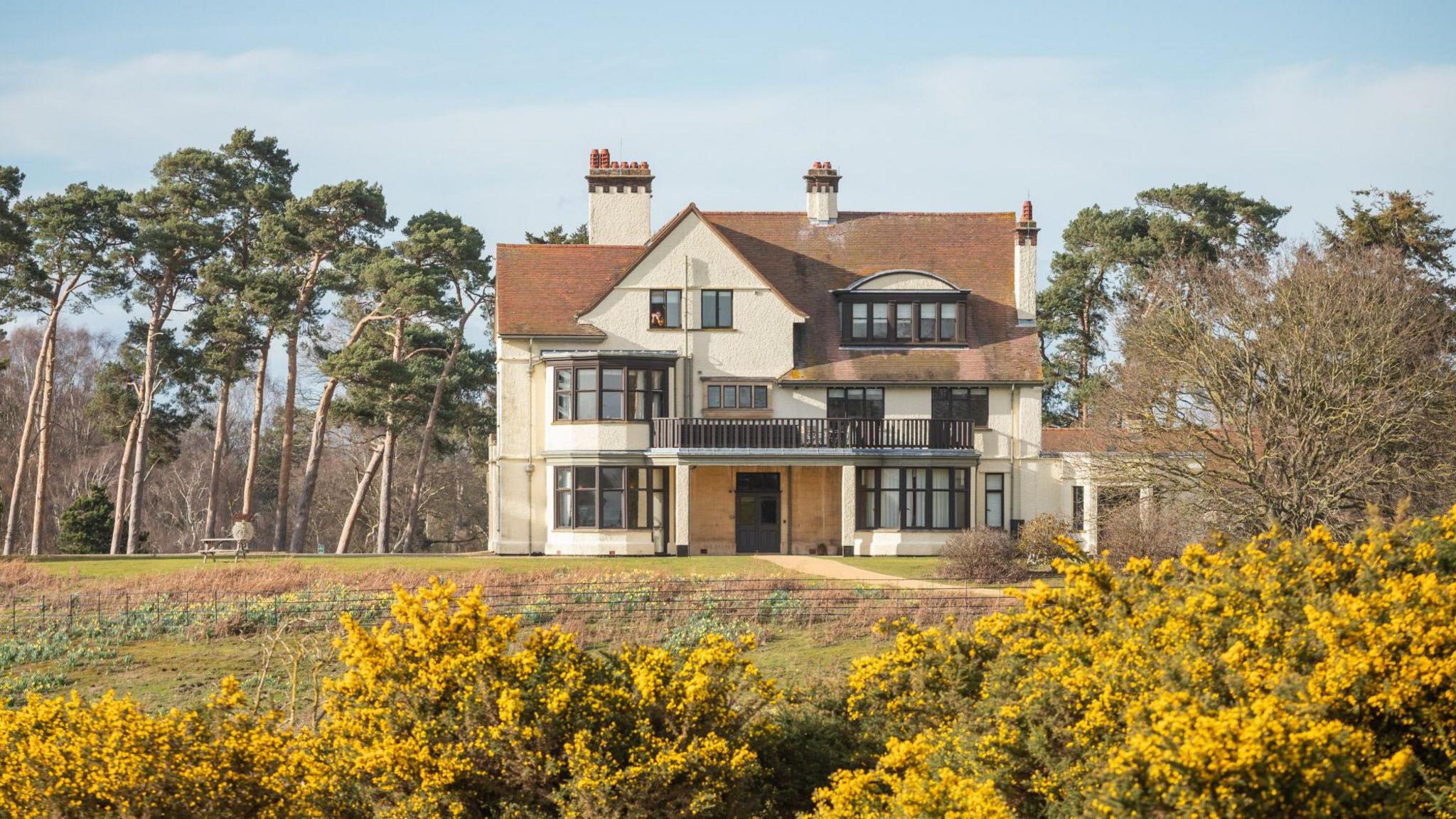 A view of the exterior of Tranmer House at Sutton Hoo. It is a largely white house with a brown roof. It is surrounded by trees and gorse and the front garden slopes away into a hollow.