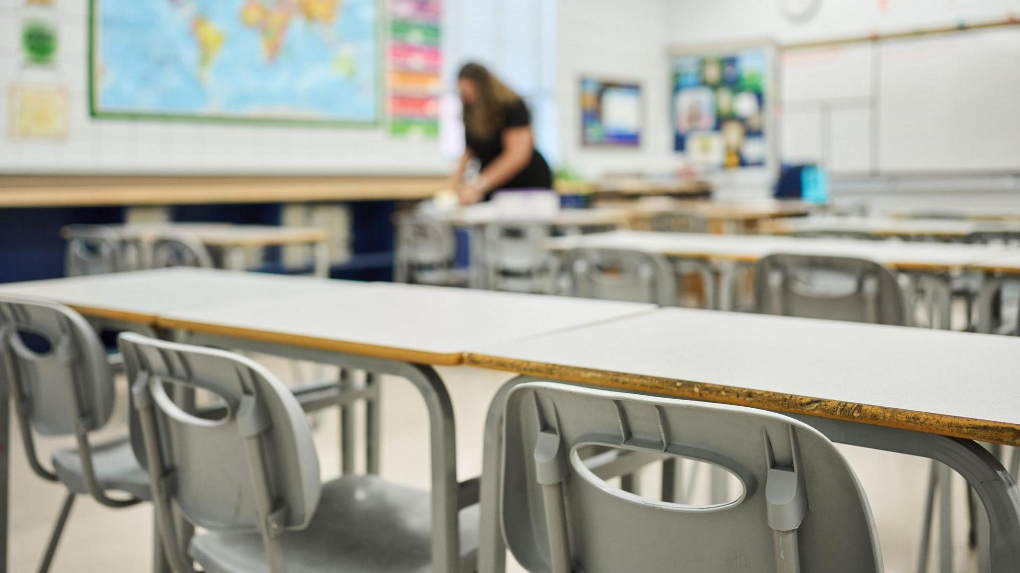 Grey chairs at desks sitting in rows in the classroom of a school with a female teacher working in the background. She is wearing a black topped and is blurred. There is a map of the world on the wall.