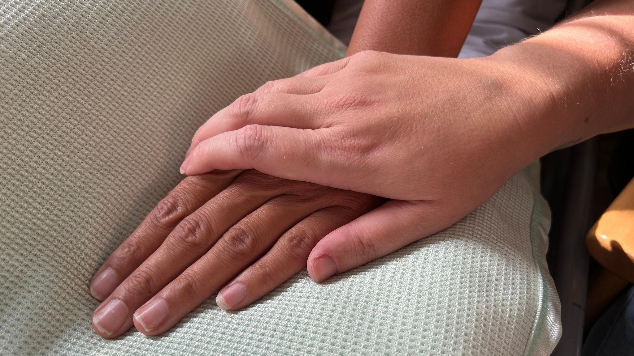 A close up of a hand in a lap with another hand laid on top of it for support. A hospital light coloured gown can be seen underneath the hands.