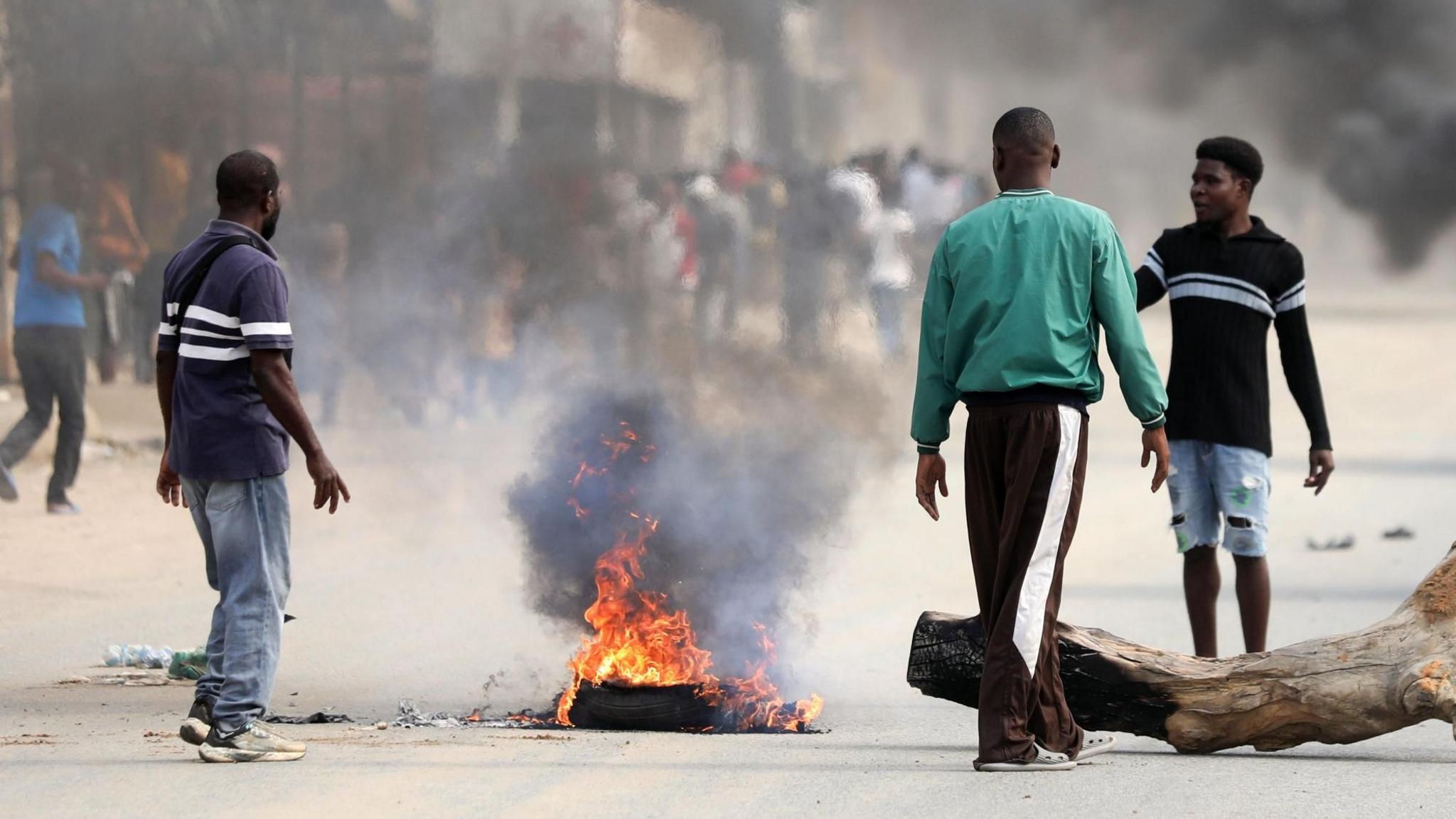 Three men burn wood on a road in Luanda. Next to one of the men is a partially burnt tree trunk lying on the ground.