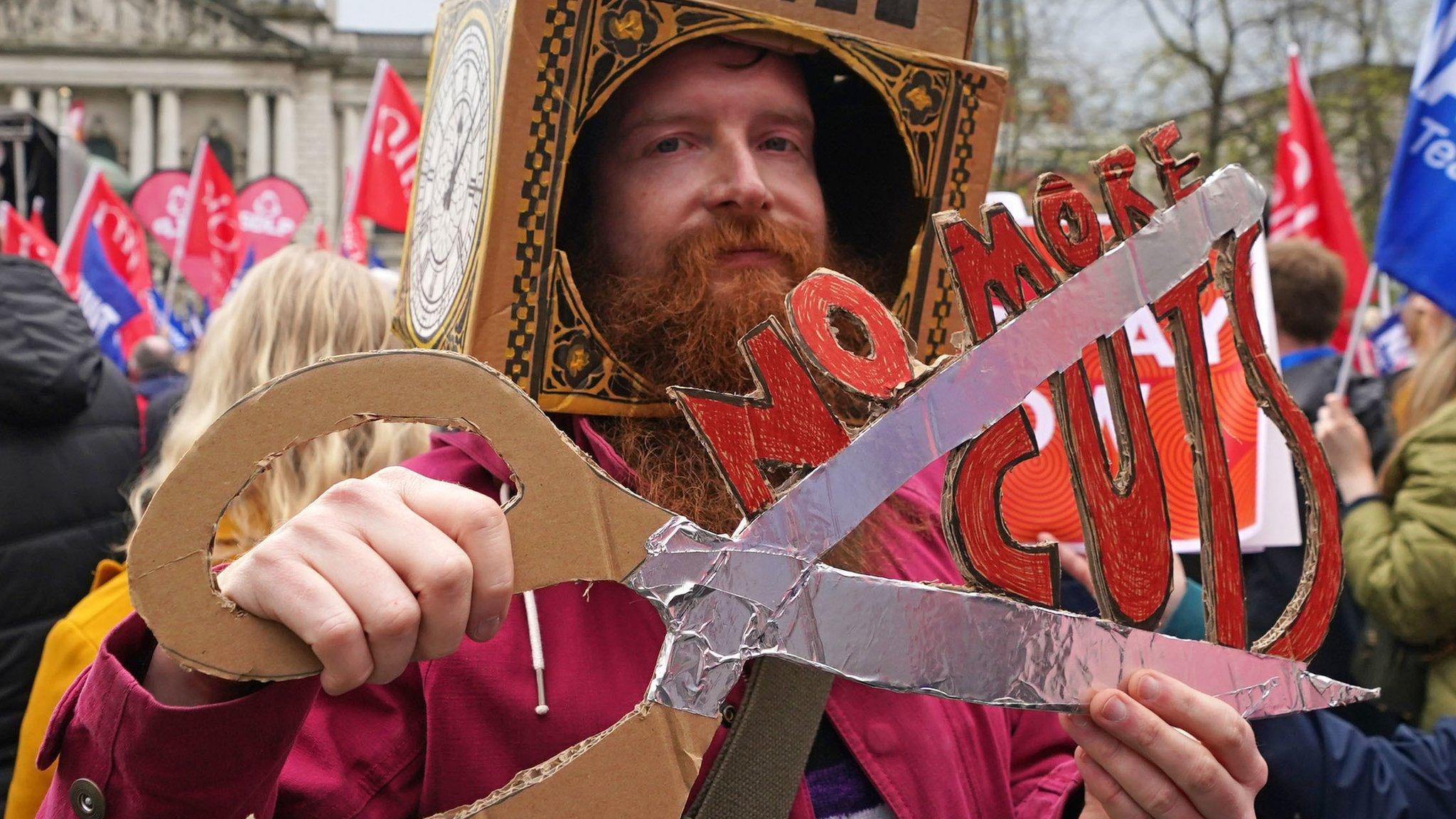 A man holds a cardboard placard that reads: NO MORE CUTS