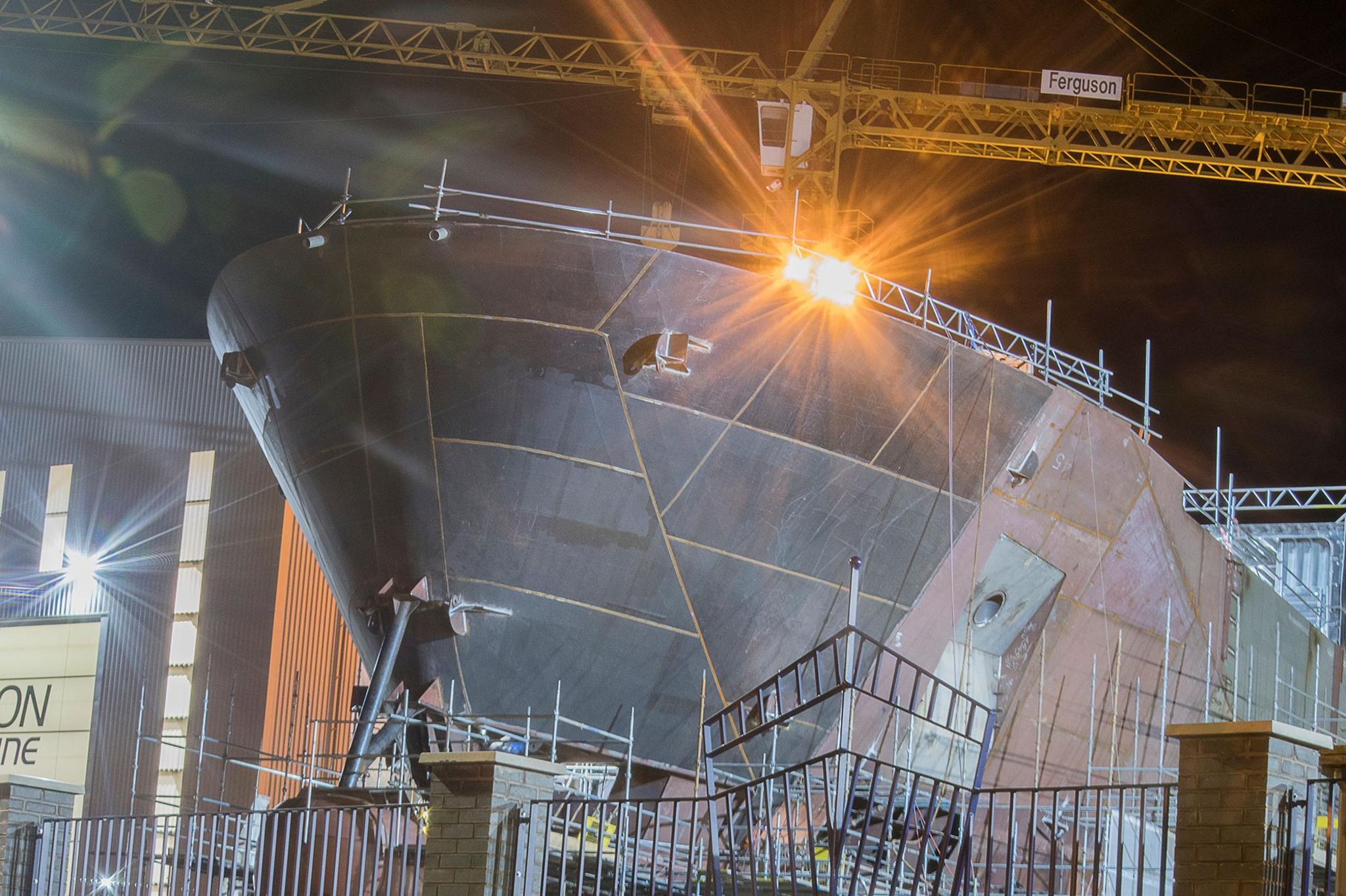 A unpainted ship being constructed on a slipway, with a yellow crane in the background