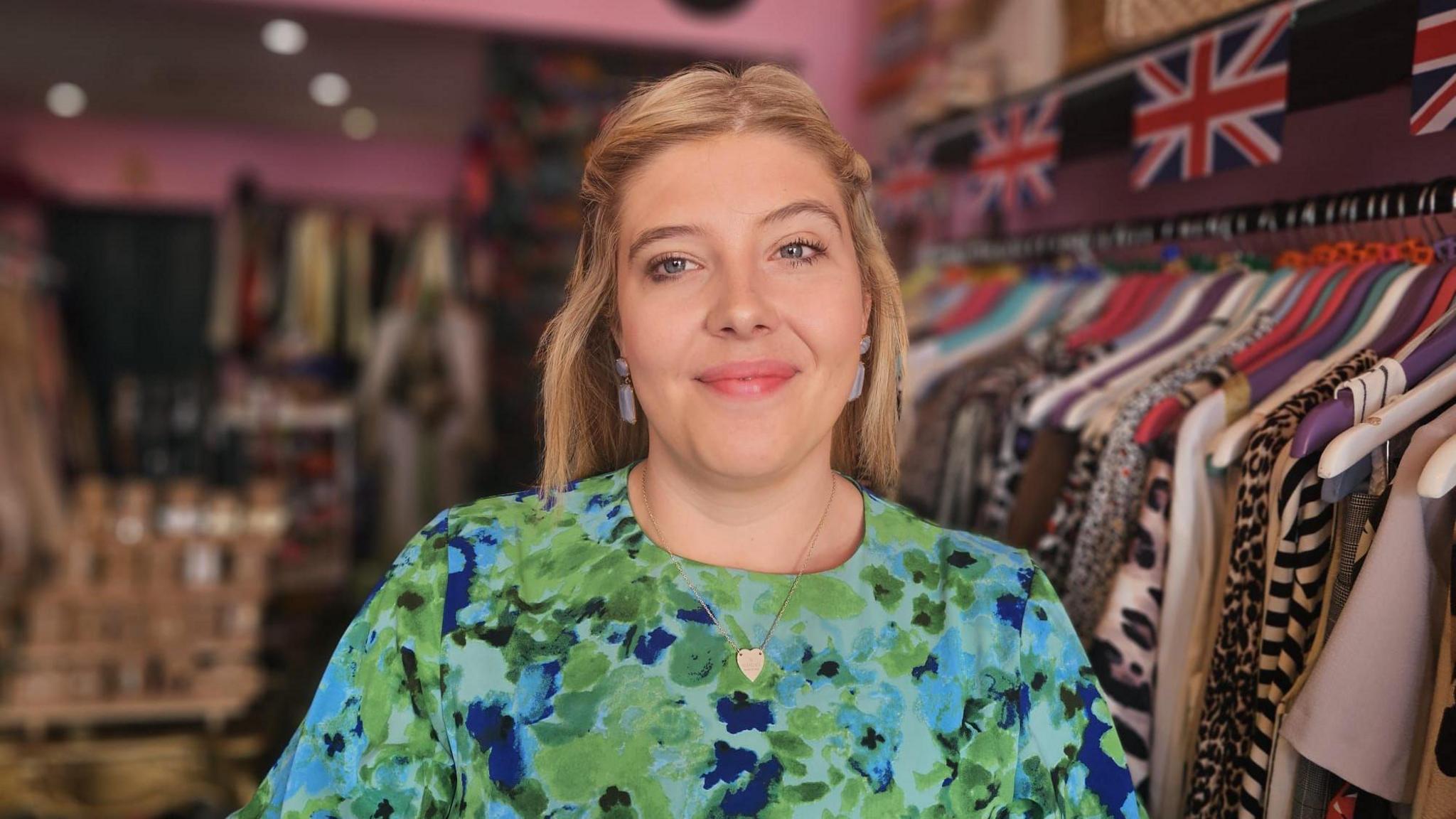 Tilly wears a green and blue floral dress and is smiling at the camera. She had a heart shaped silver necklace on and light blue dangly ear rings. Her hair is blonde and shoulder length, it's down but clipped off her face. Behind her is a rack of clothing and union jack bunting.