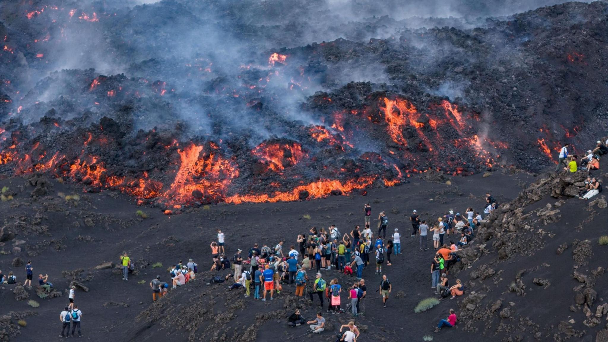People gather to watch as lava flows down Mount Etna during a period of heightened volcanic activity, near Catania, Sicily, southern Italy, 13 August 2026. 