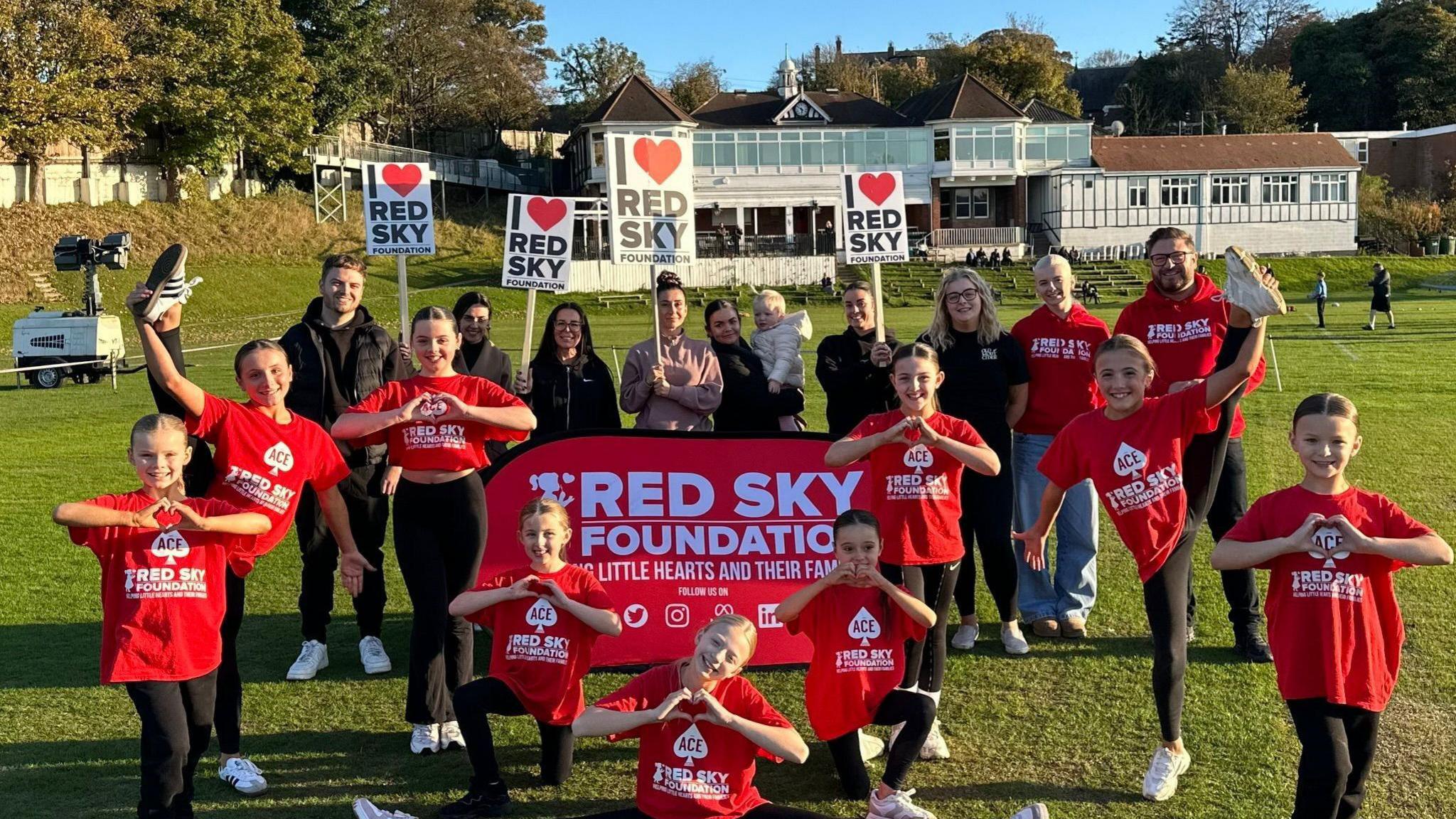 Children wearing red tops stand around a banner reading Red Sky Foundation. They are on the grounds of Ashbrooke Sports Centre in Sunderland with the pavilion behind