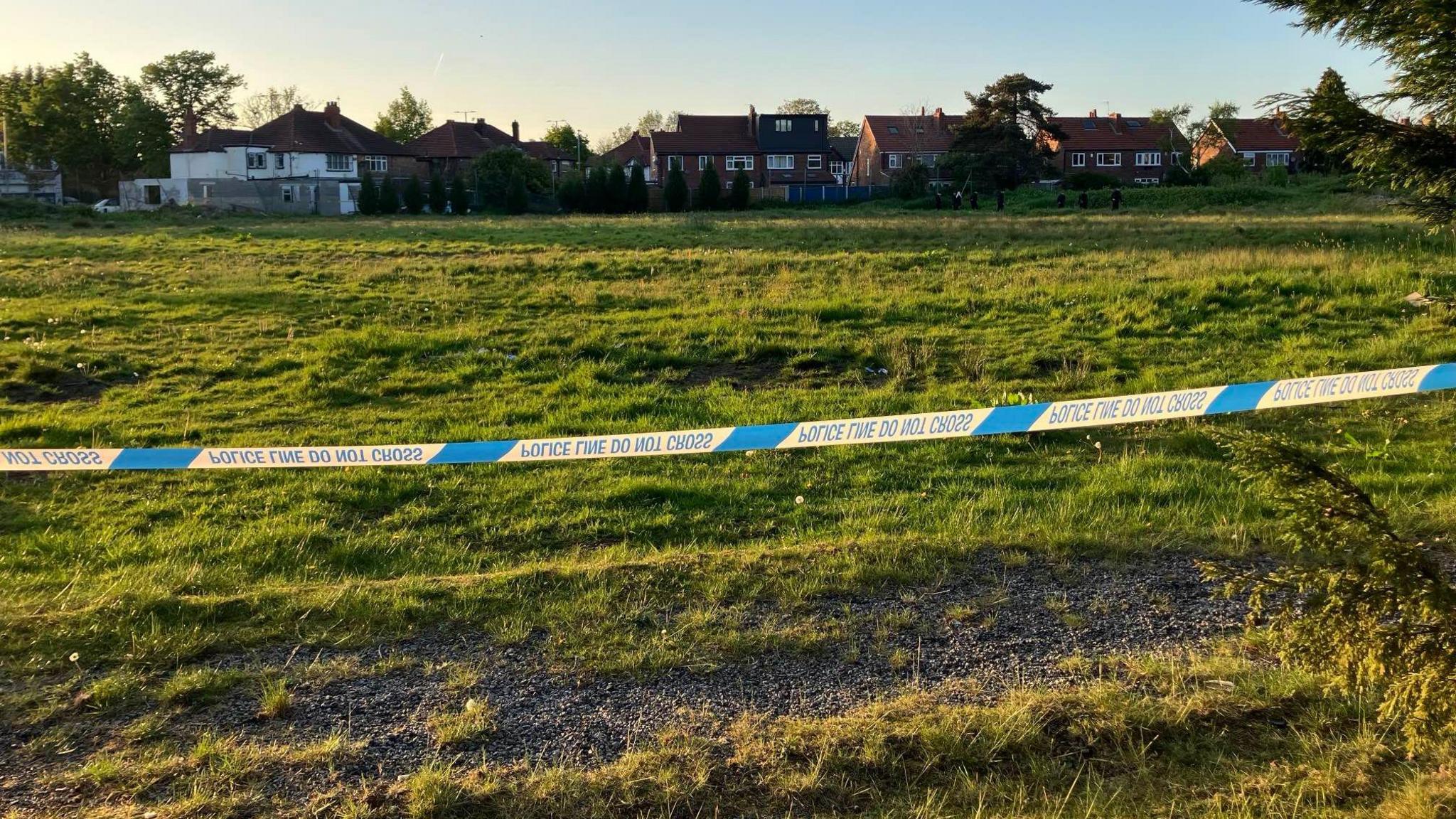 Former cricket field is cordoned off with blue and white police tape - there are houses in the distance
