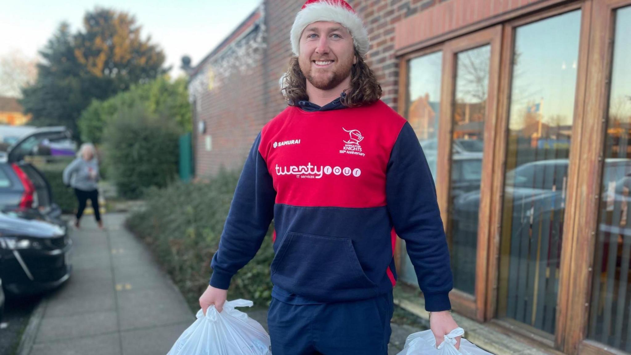 Connor Davidson has long curly hair and is wearing dark blue and red sports clothes and a Santa hat. He is smiling as he lifts two white shopping bags.