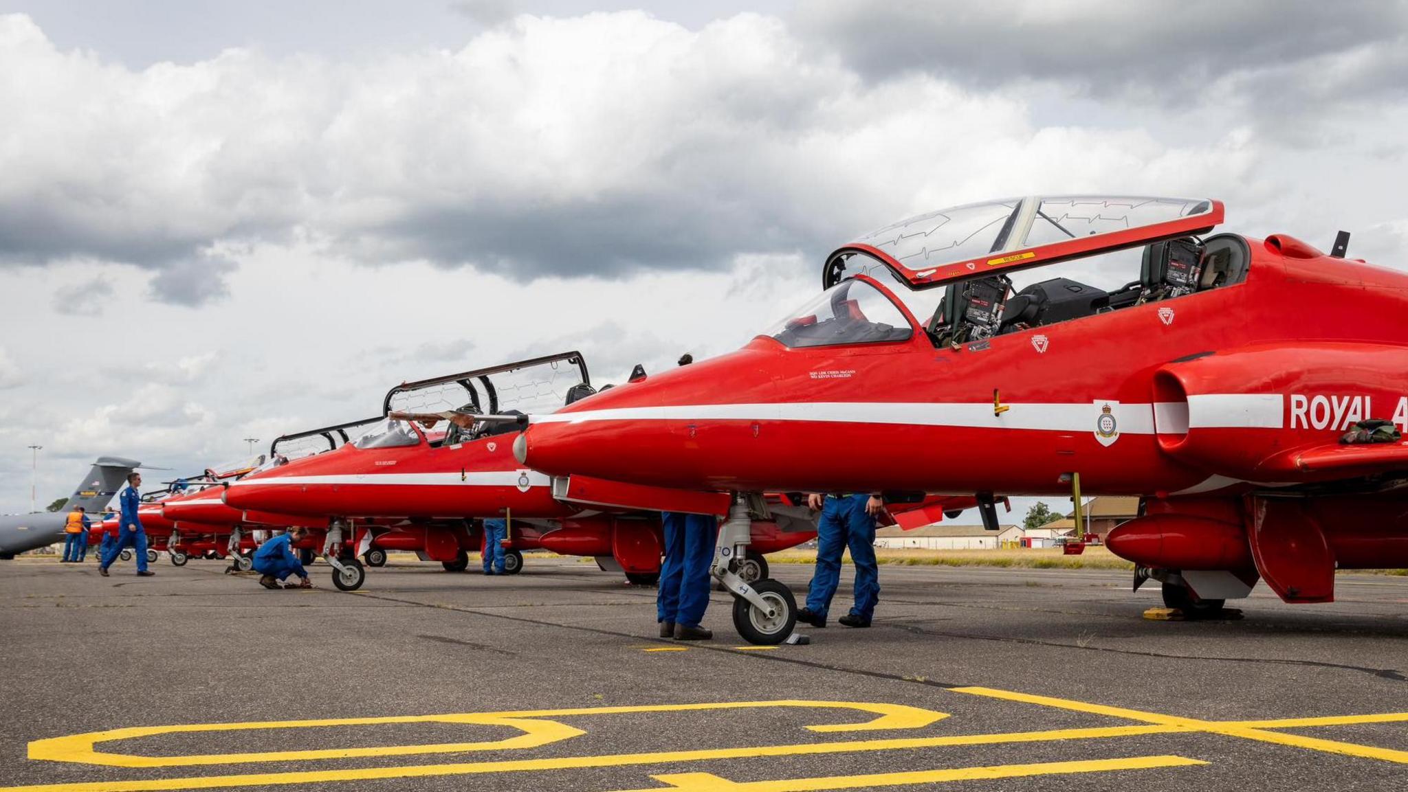 A number of red planes are lined up on a concrete runway. There are several men in blue overalls checking over the planes