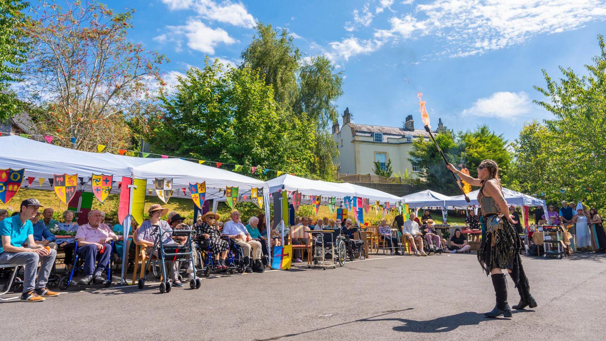 A woman holding a flaming stick stands in front of a row of care home residents who are sitting down under gazebos in the sunshine