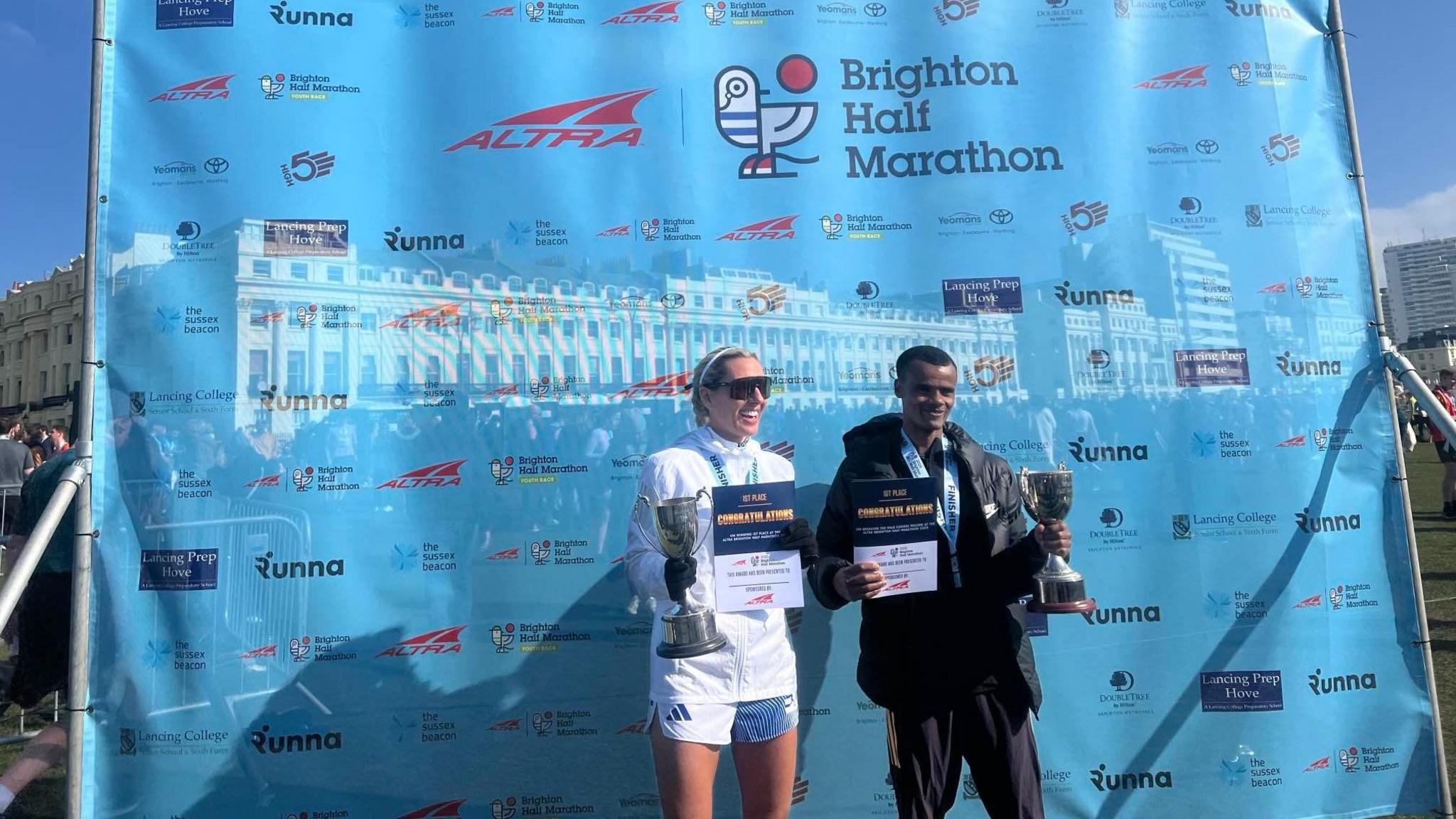 The winners of the men's and women's Brighton Half Marathon event stand with their trophies in front of a pale blue plastic sheet with the sponsors' names across it.