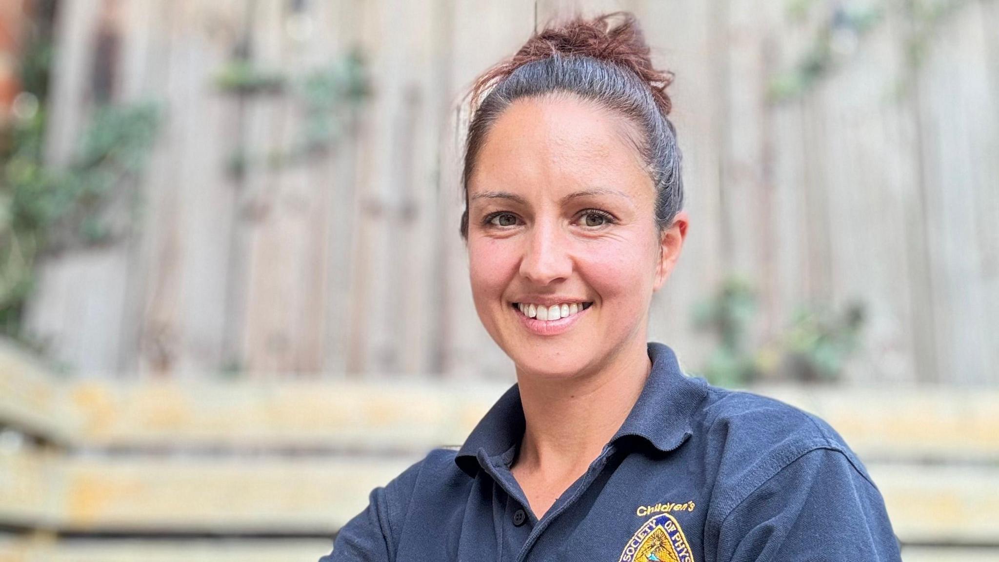A woman is smiling into the camera. She's sitting outdoors. Her hair is tied back and she's wearing a t-shirt.