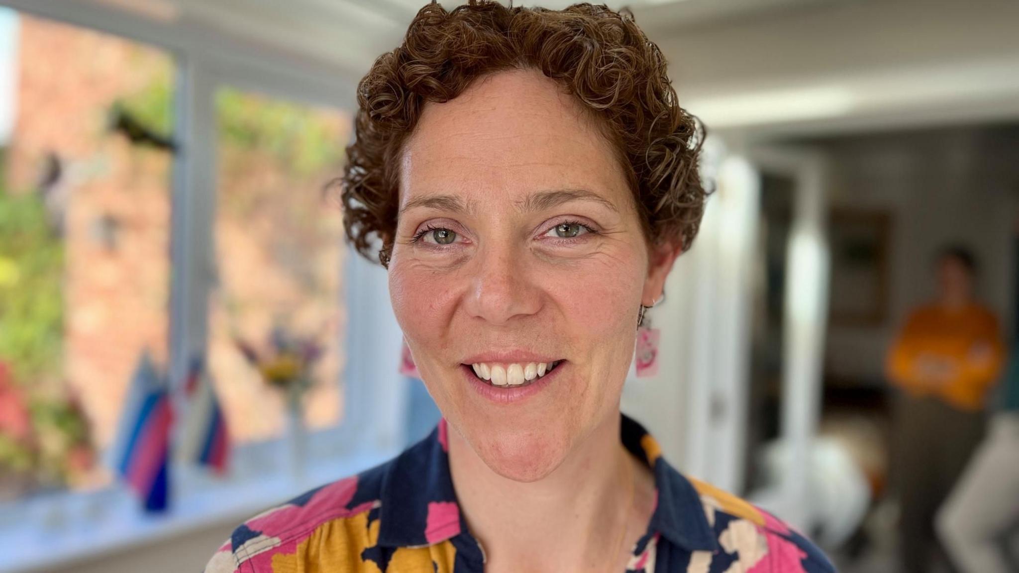 Julia Beeden looks at camera smiling, she has pink square earrings in and a flowery top with pink and orange flowers, she has short, curly hair after having cancer treatment and is standing in her kitchen in front of a window