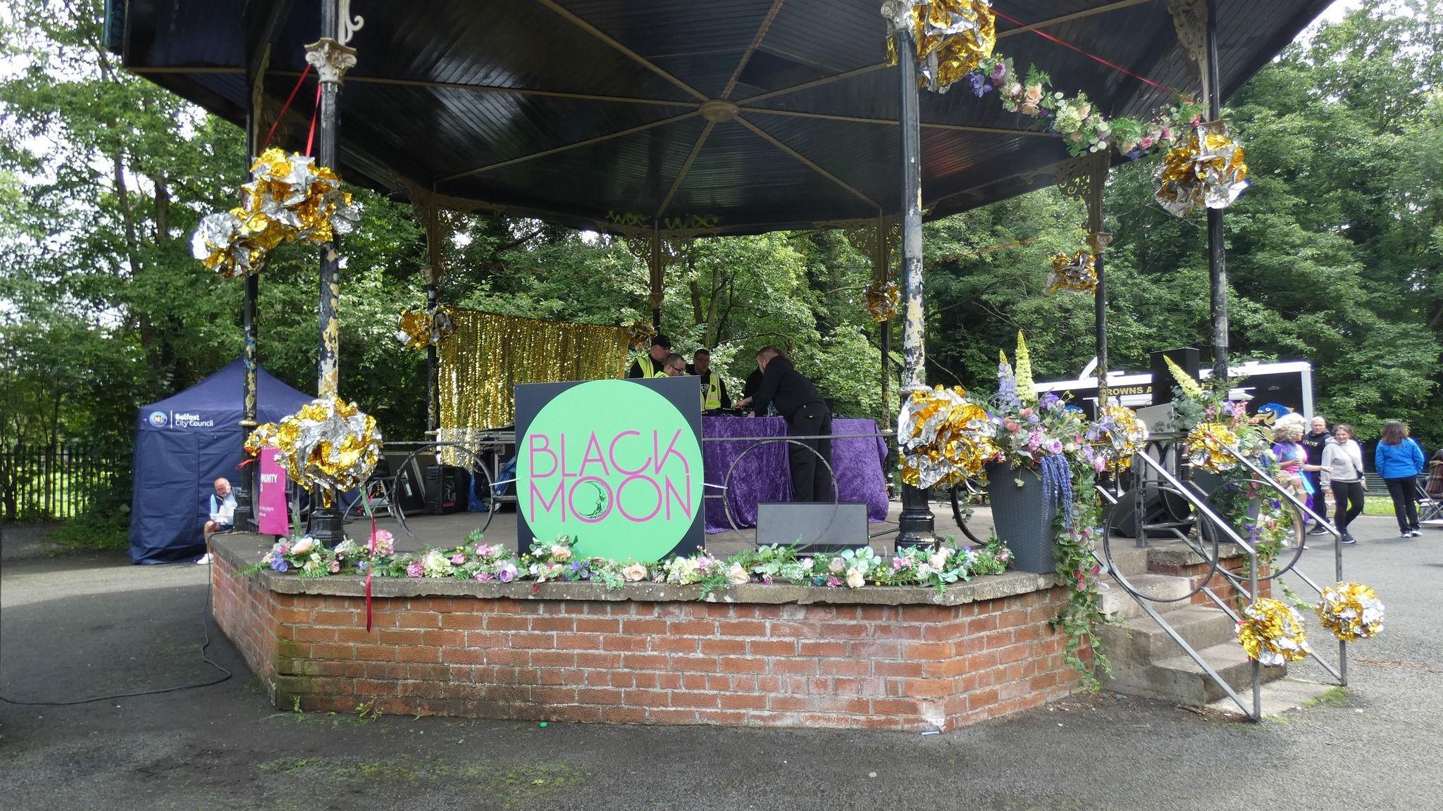 A red brick bandstand has black posts separating it from its black roof. There is a large circular badge which has the words 'Black Moon' written on it. Behind the stand is a navy tent. There are yellow floral arrangements all over the bands railings. As well as a table with a purple table cloth in the centre.