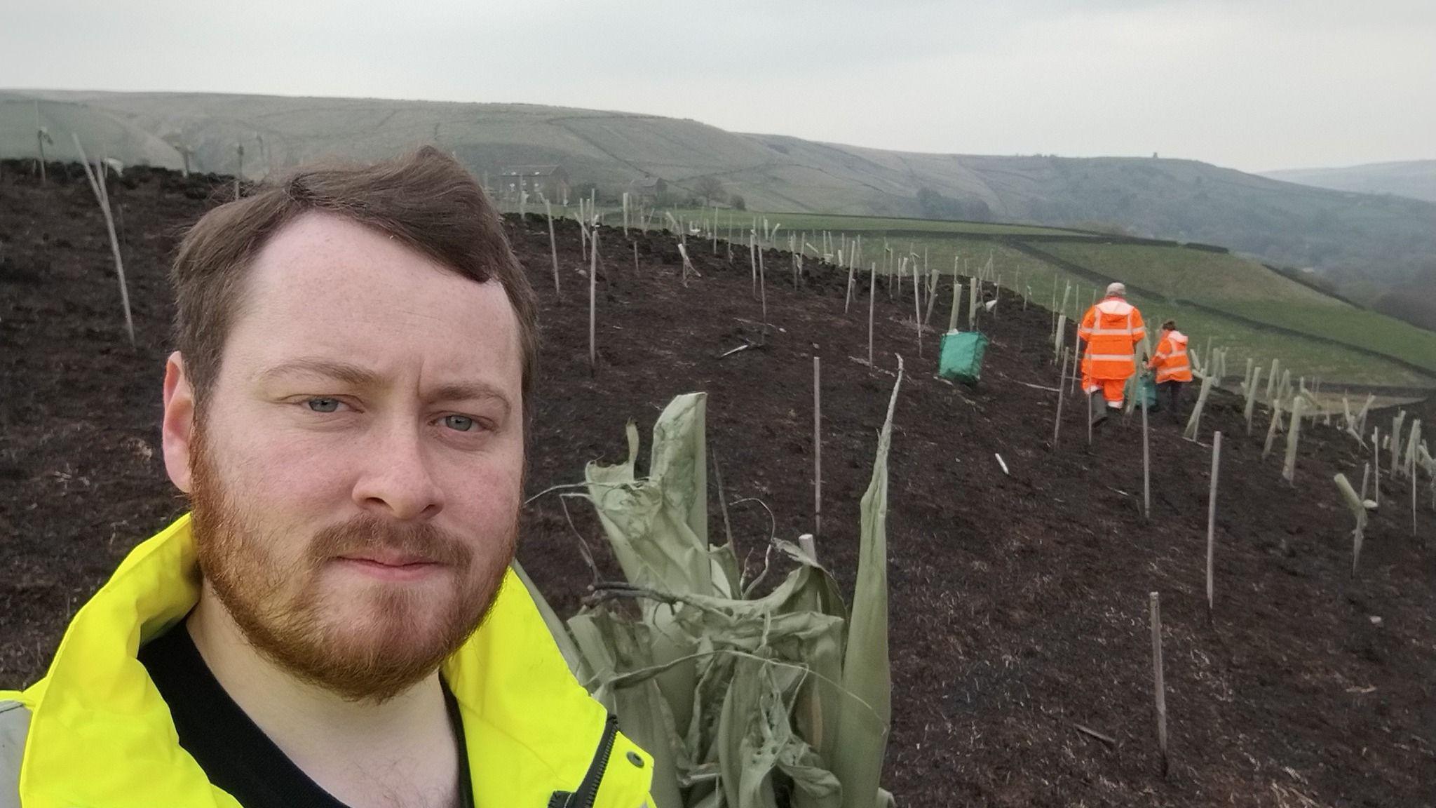 Matthew McLoughlin standing on moorland with a heap of burnt plastic tree cylinders behind him. He is wearing a black T-shirt and a yellow hi-vis jacket. There are two people in orange hi-vis jackets in the distance. The sky is grey and cloudy.