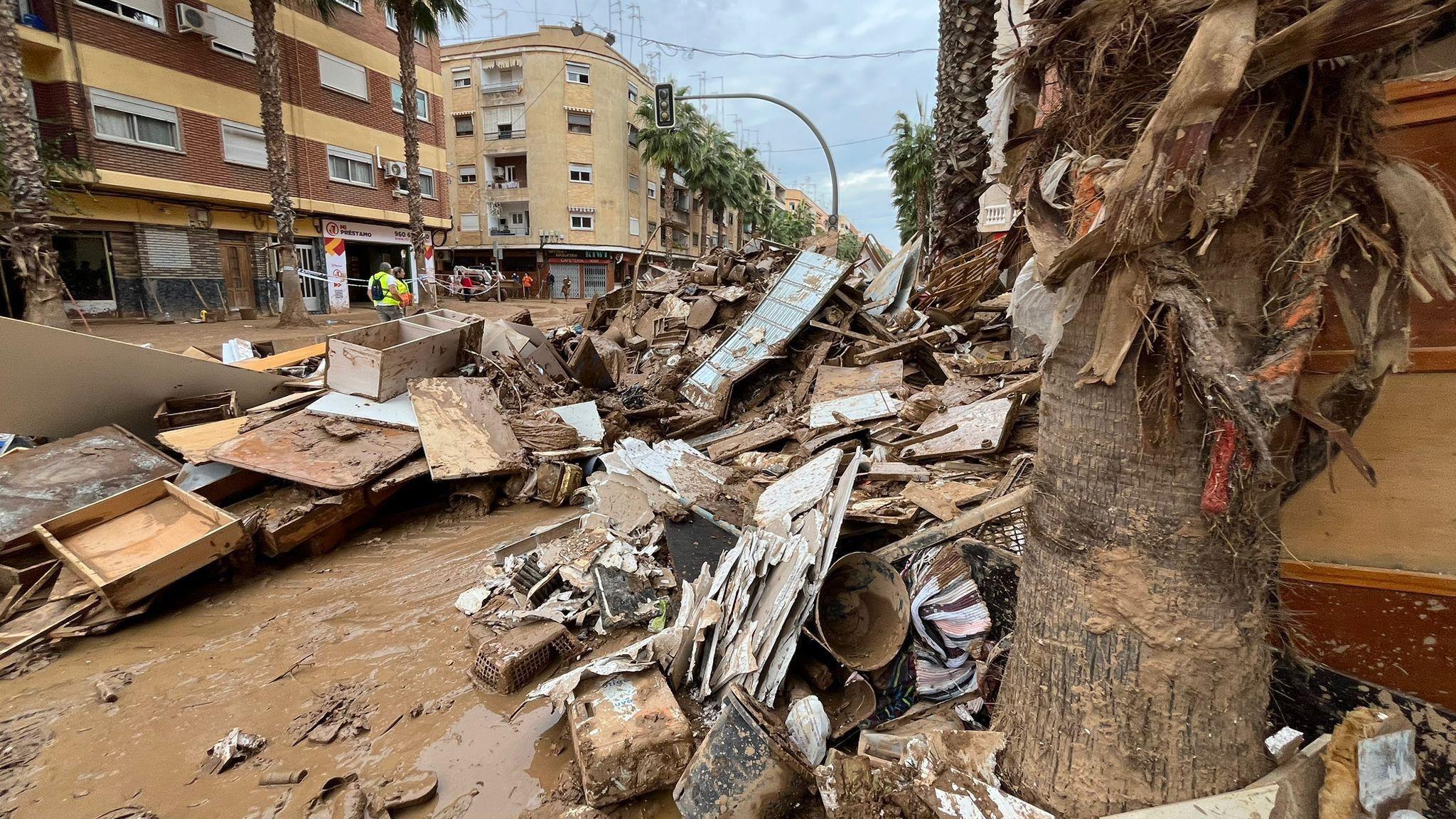 A street in Valencia covered in debris following the flash floods. The ground is muddy and still wet. There is furniture, parts of buildings and plasterboard strewn across the road in large stacked up piles.