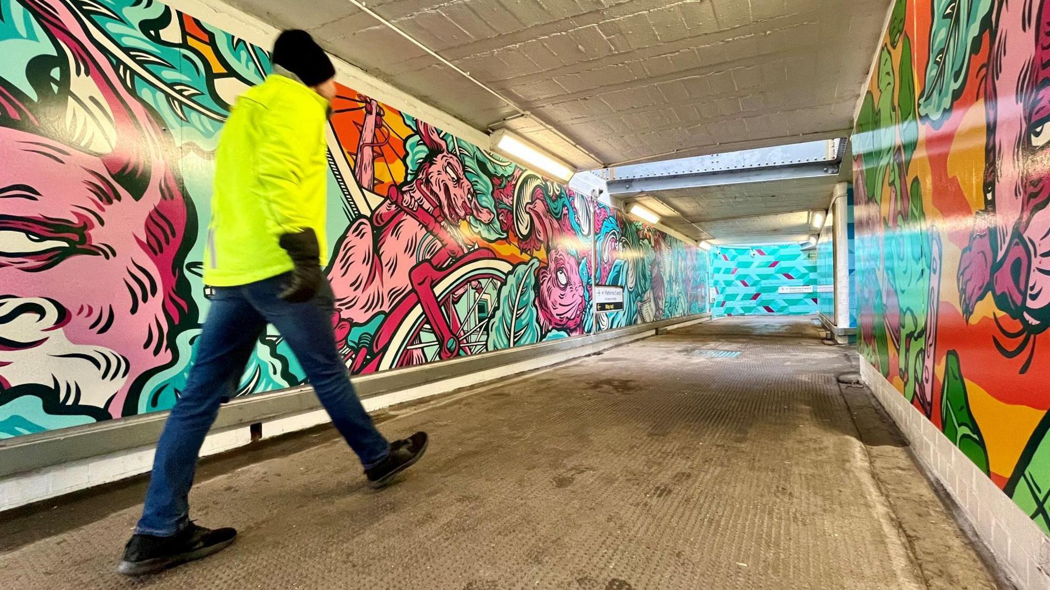 A man in a bright yellow high-vis jacket, black bobble hat and blue jeans walks through an underpass at Bedminster Station in Bristol. All the walls around him have colourful cartoon-style murals on them