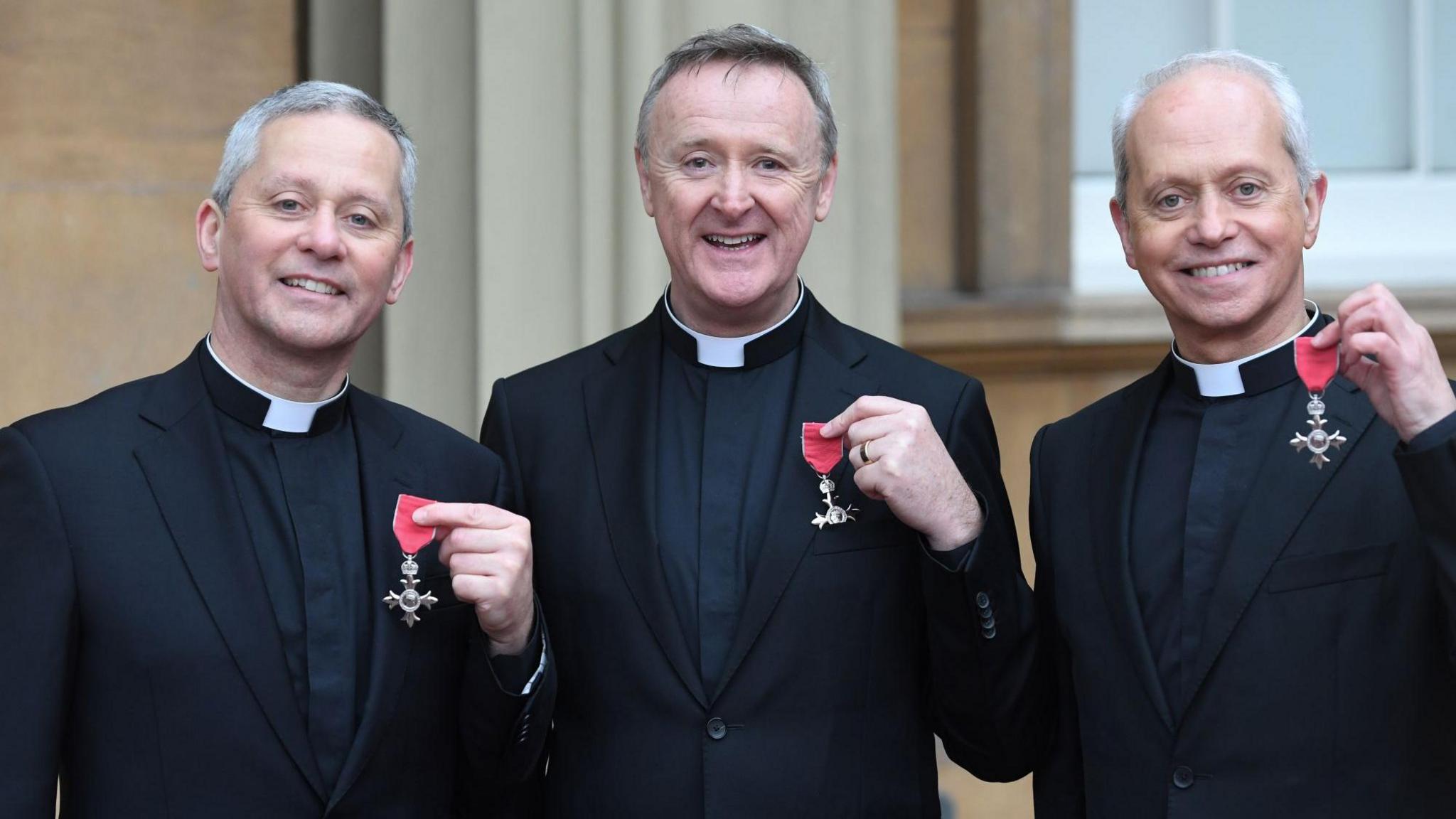 Three priests - Fr Martin O'Hagan, Fr David Delargy and Fr Eugene O'Hagan hold up their MBE awards while smiling for the camera outside of Buckingham Palace. They wearing black priest vestments and collars.