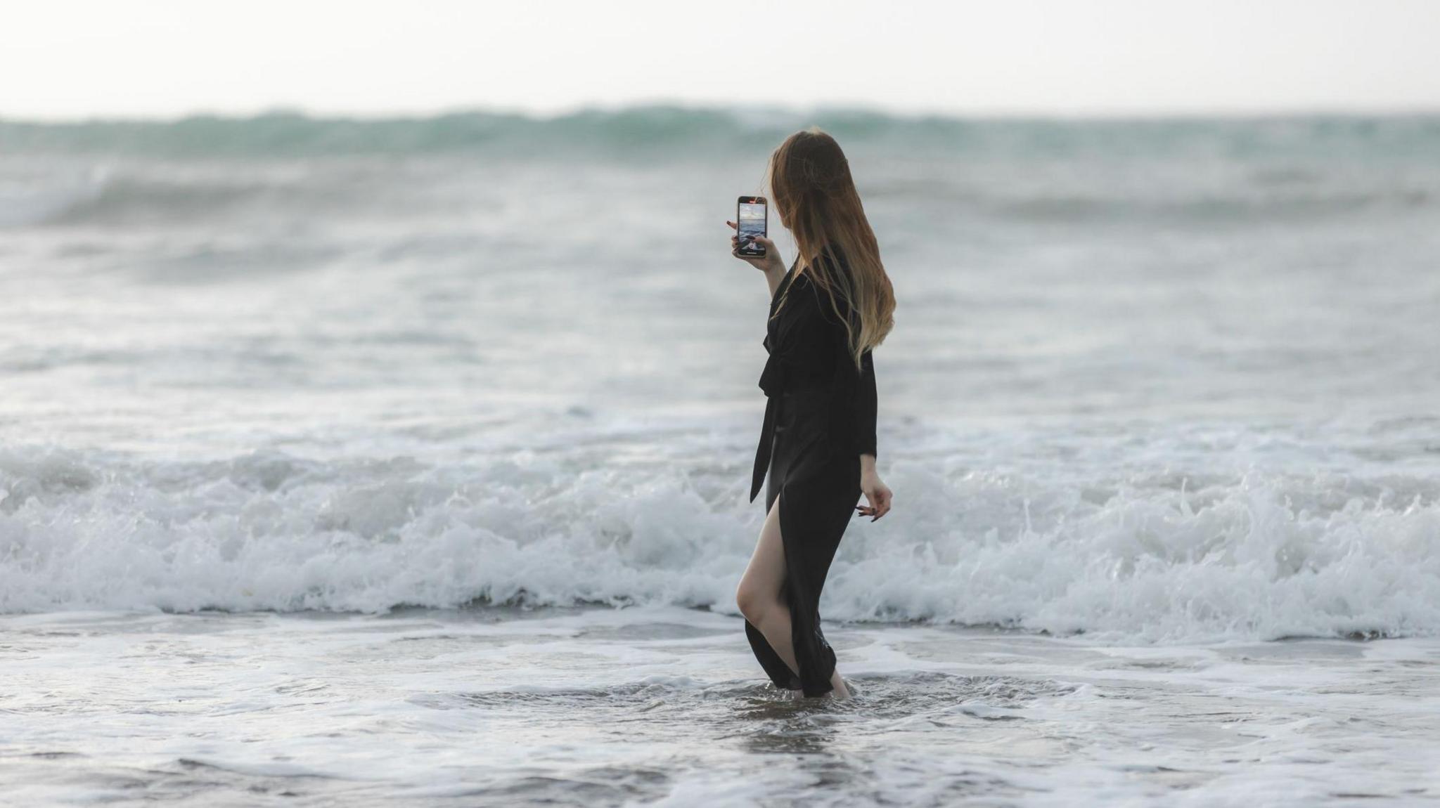 A woman with long dark blonde hair stands in the ocean. She is facing away from the camera and holding her phone in her right hand, she is recording the view of the ocean waves. The woman wears a long sleeved black dress, which has a long side split in the skirt showing her left leg.